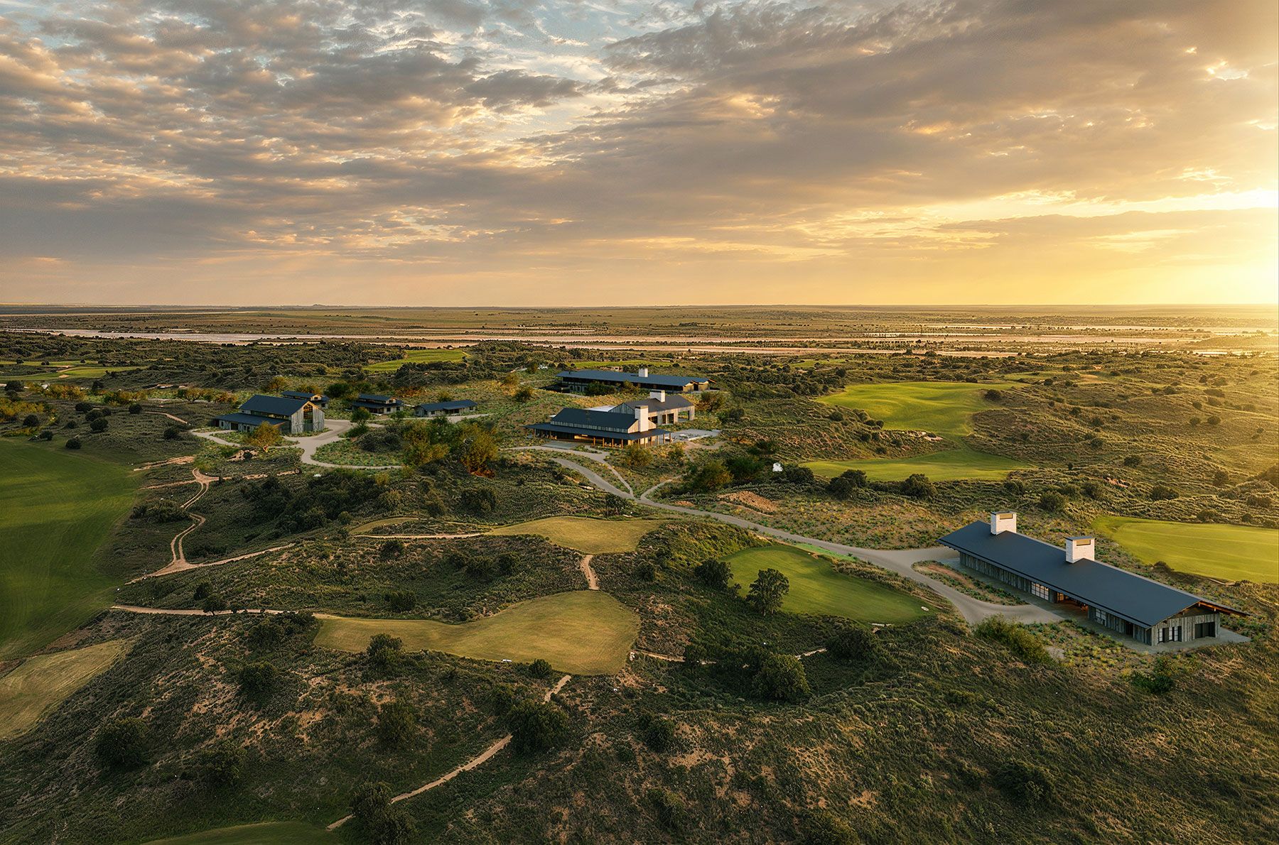 Aerial view of a luxury golf complex and modern wooden resort buildings spread across rolling green hills at sunset.