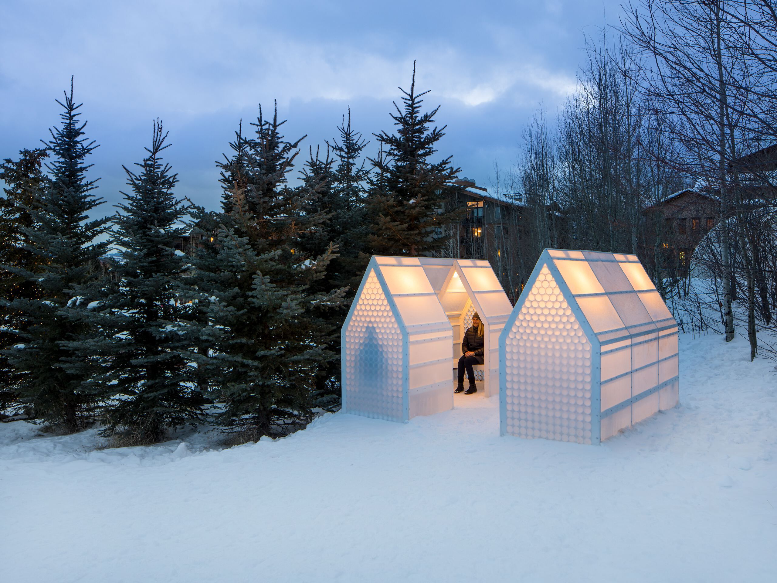 Three glowing, house-shaped winter shelters with translucent walls made of stacked circular patterns sit in a snow-covered landscape at dusk. A woman bundled in a winter coat sits inside the central hut, which emanates a warm light. Evergreen trees flank the left, while bare trees and distant buildings are visible on the right under a blue-grey sky.
