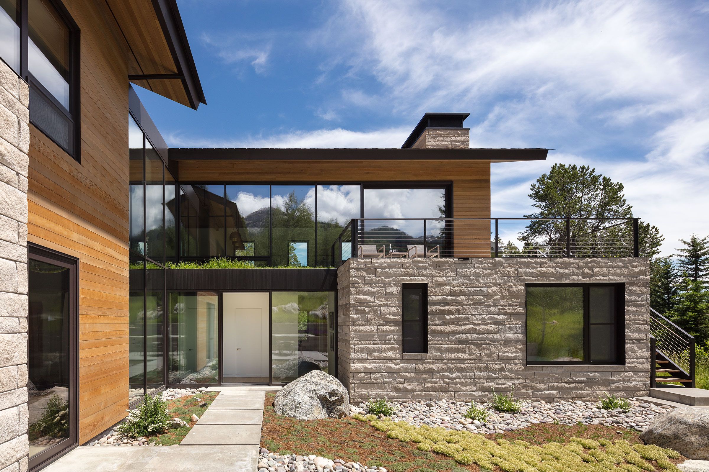 Exterior view of a contemporary two-story house combining light stone masonry and warm wood siding, with large glass windows reflecting a blue sky with clouds.