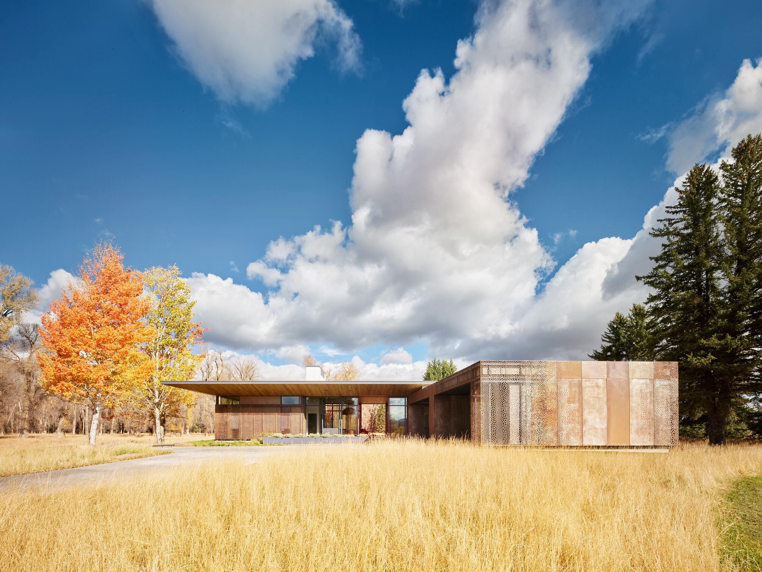 Modern residence with weathered Corten steel and perforated screens, featuring expansive glass walls, set amidst a golden grass field, vibrant orange and yellow autumn trees, and tall evergreens under a bright blue sky with fluffy white clouds.