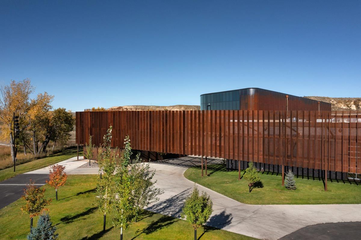 A striking modern building features a facade of rusty brown vertical panels, with a curved glass section visible at one end, all beneath a clear blue sky. The elevated structure casts shadows over green lawns, autumn trees, and a winding paved path in the foreground, while hills frame the distant background.