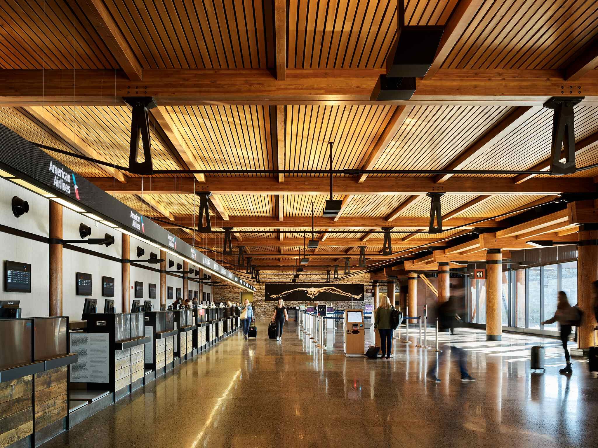Modern airport check-in hall featuring a high wooden beam and slat ceiling, polished terrazzo floor, and rustic wooden columns. Rows of airline check-in counters, including American Airlines and Delta, line the left. Travelers with luggage move through the spacious area, with some motion blur. A large mural of an eagle flying over mountains is visible on the far wall.