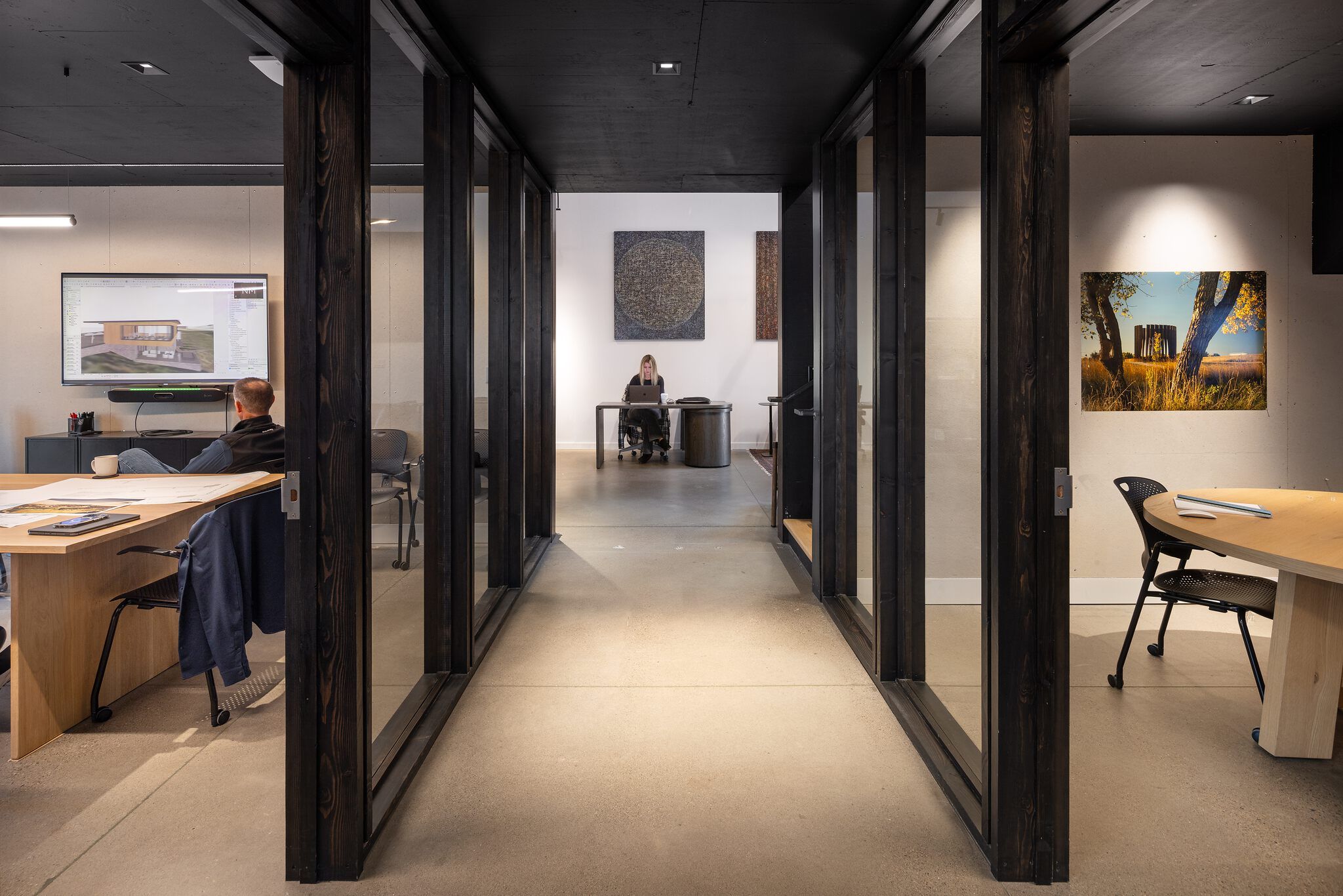 Modern office interior with a central dark-framed hallway and glass partitions. A man works on architectural plans at a large monitor on the left, while a woman works on a laptop at a desk at the hallway's end. A meeting table is visible in an adjacent room on the right. The space features dark wood accents, concrete floors, and curated artwork.