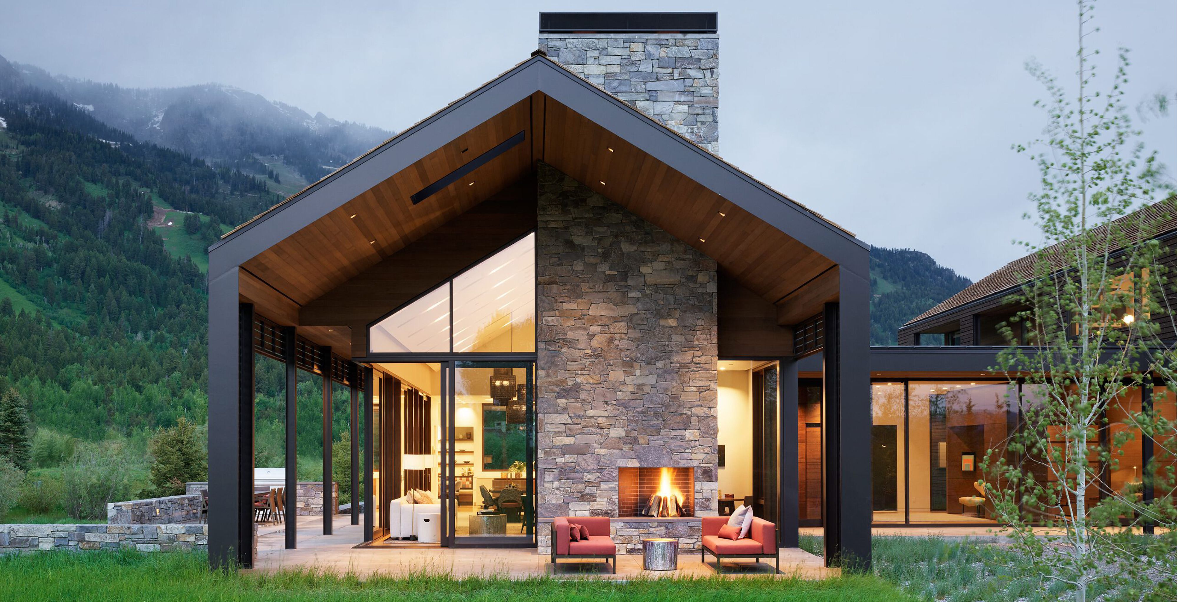 Contemporary mountain home with a stone and floor-to-ceiling glass exterior, featuring a covered patio with an outdoor fireplace and lounge seating. Set against a backdrop of lush green mountains at dusk.