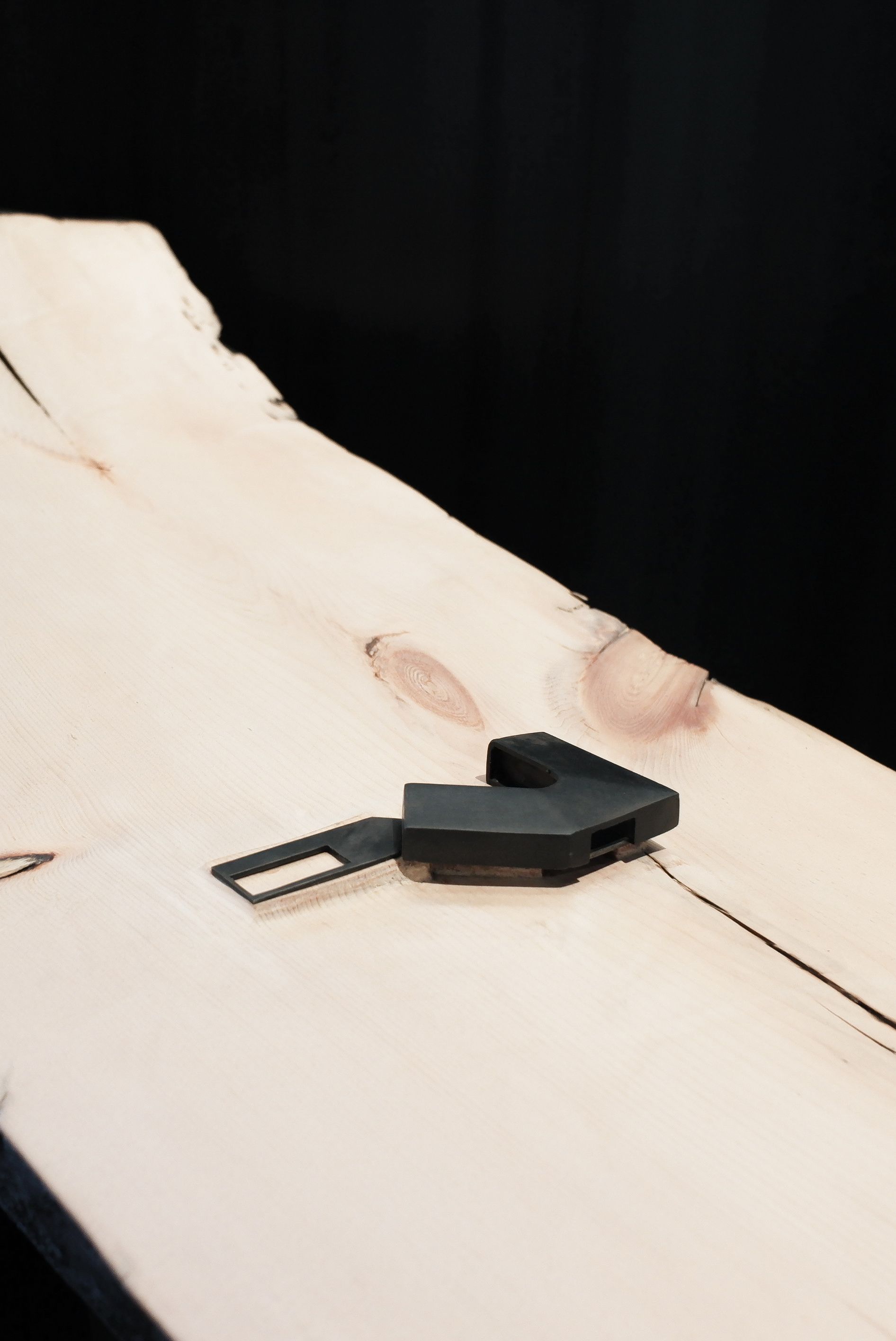 A light-toned live edge wood slab table with visible grain and natural cracks, featuring a black, angular seatbelt buckle release tool on its surface. The table is set against a dark background.