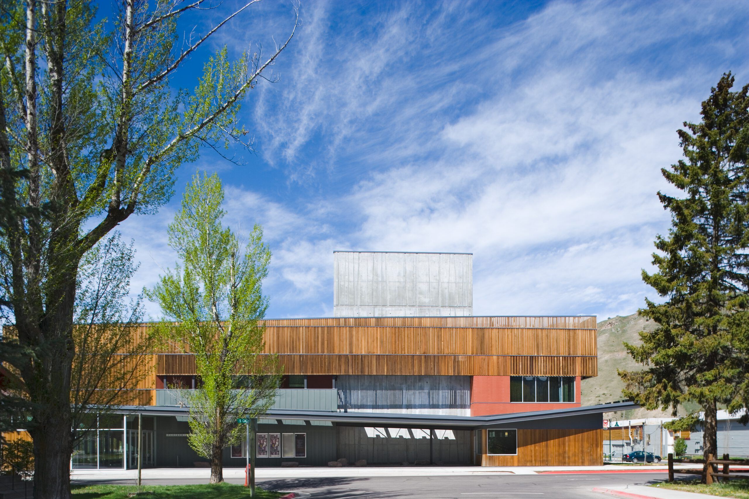 Modern performance arts center with a facade of vertical wood paneling, gray concrete, and reddish accents, featuring an angular entrance canopy. Lush green trees frame the building under a bright blue sky with wispy white clouds, with distant mountains visible in the background. A street sign for S Cache is near the entrance.