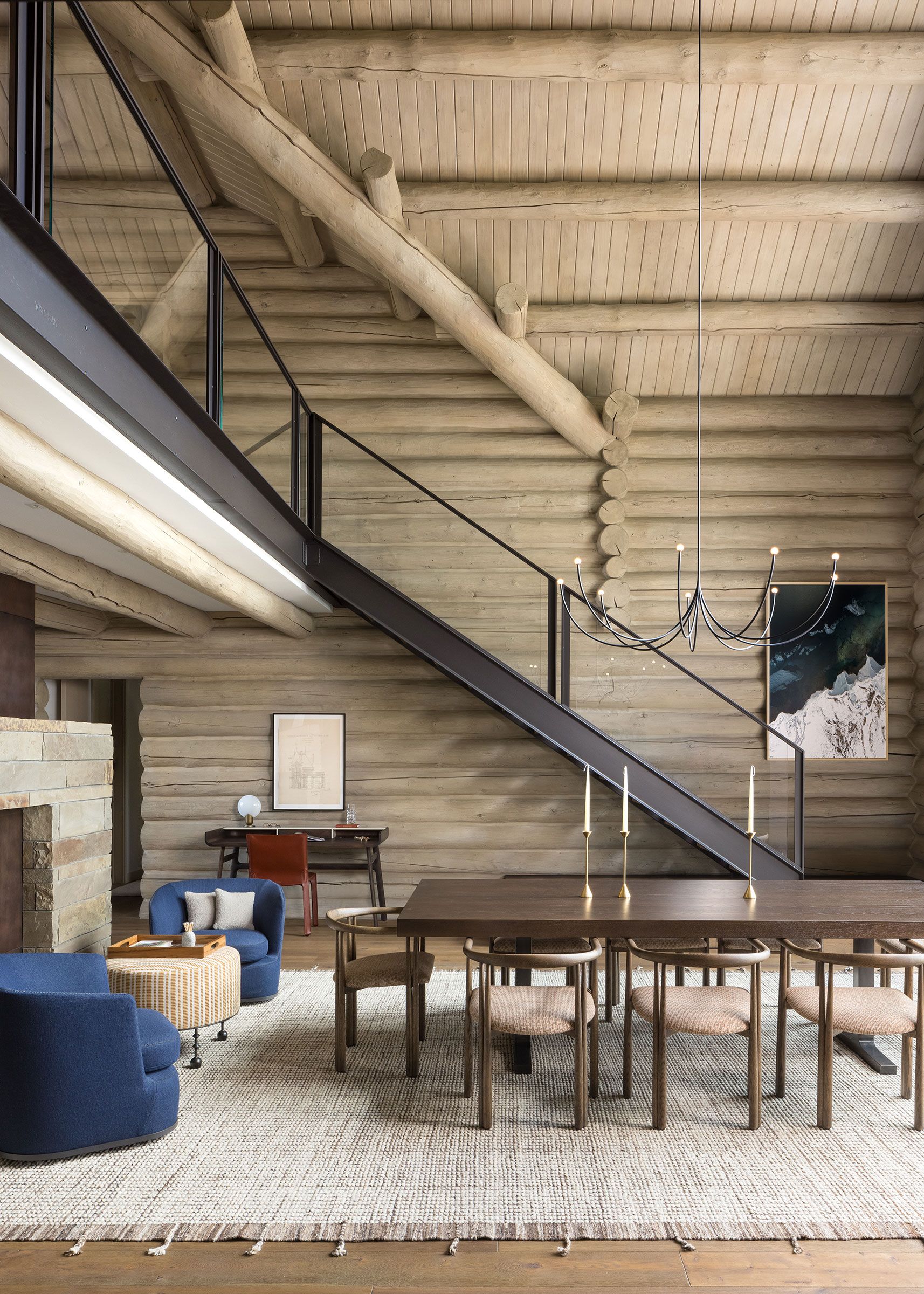 Spacious, open-concept log cabin interior featuring a long wooden dining table, modern chandelier, blue armchairs, and a sleek metal staircase leading to a loft.