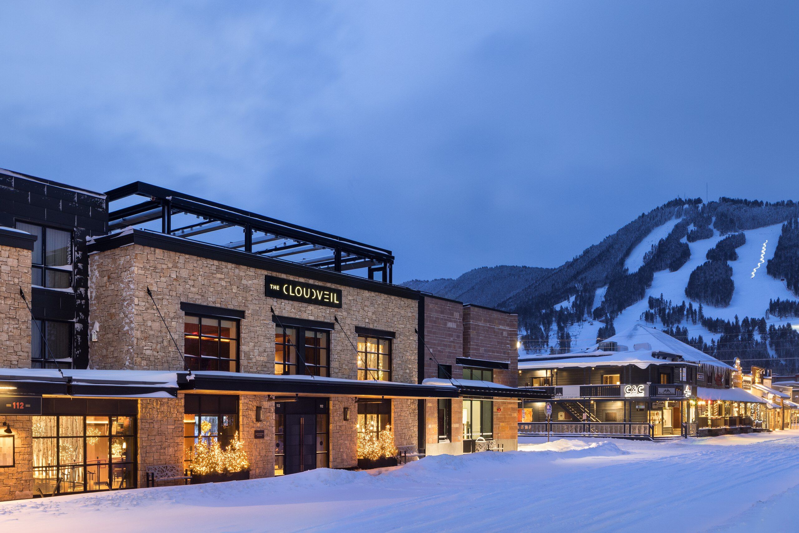 The Cloudveil hotel, featuring a stone and dark facade, is brightly lit with warm lights and decorative outdoor trees. It stands on a snow-covered street at dusk, with the snowy ski slopes of a mountain resort visible under a twilight blue sky.