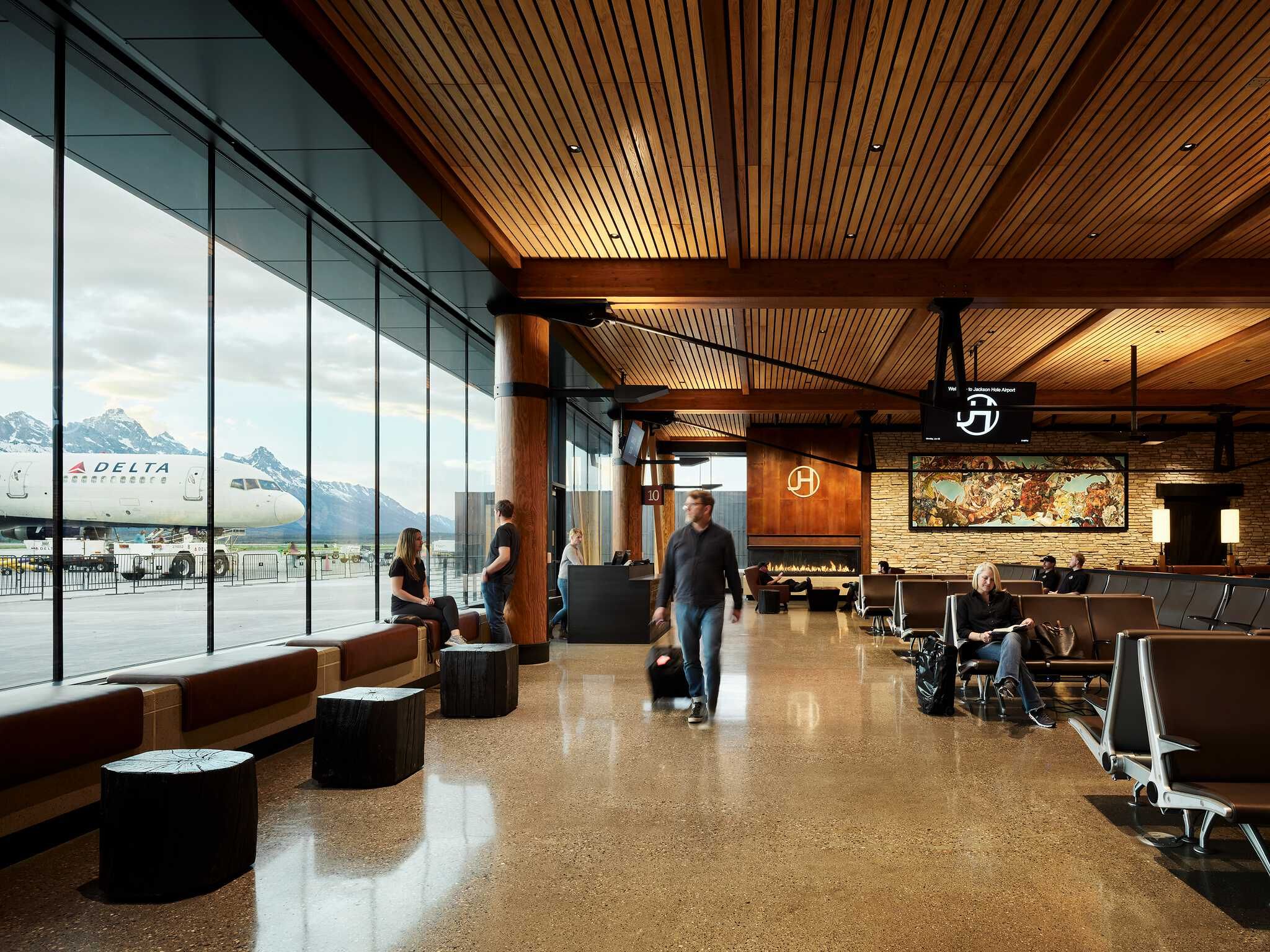 Spacious, modern airport terminal interior at Jackson Hole, featuring an expansive view of a Delta airplane and snow-capped mountains through large windows. The design blends rustic and contemporary elements with a slatted wood ceiling, exposed wooden beams, and polished concrete floors. Travelers are seated on brown benches and modern lounge chairs. A large artwork hangs above a fireplace on a stone wall, complementing the warm ambiance.