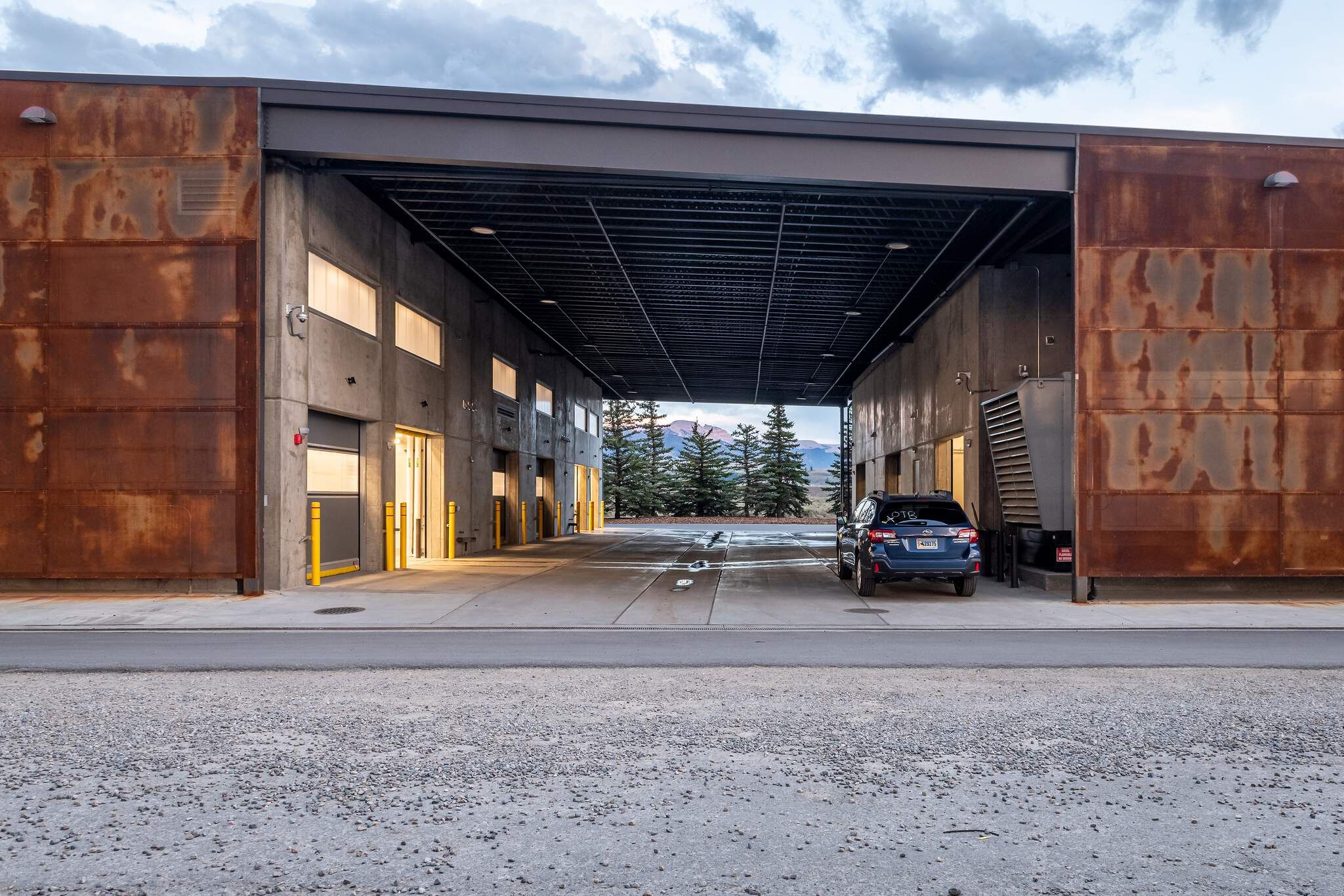 Modern building with rust-colored steel panels and a concrete open passage, where a blue SUV is parked. Beyond the passage, pine trees and a distant mountain range are visible under a cloudy sky.