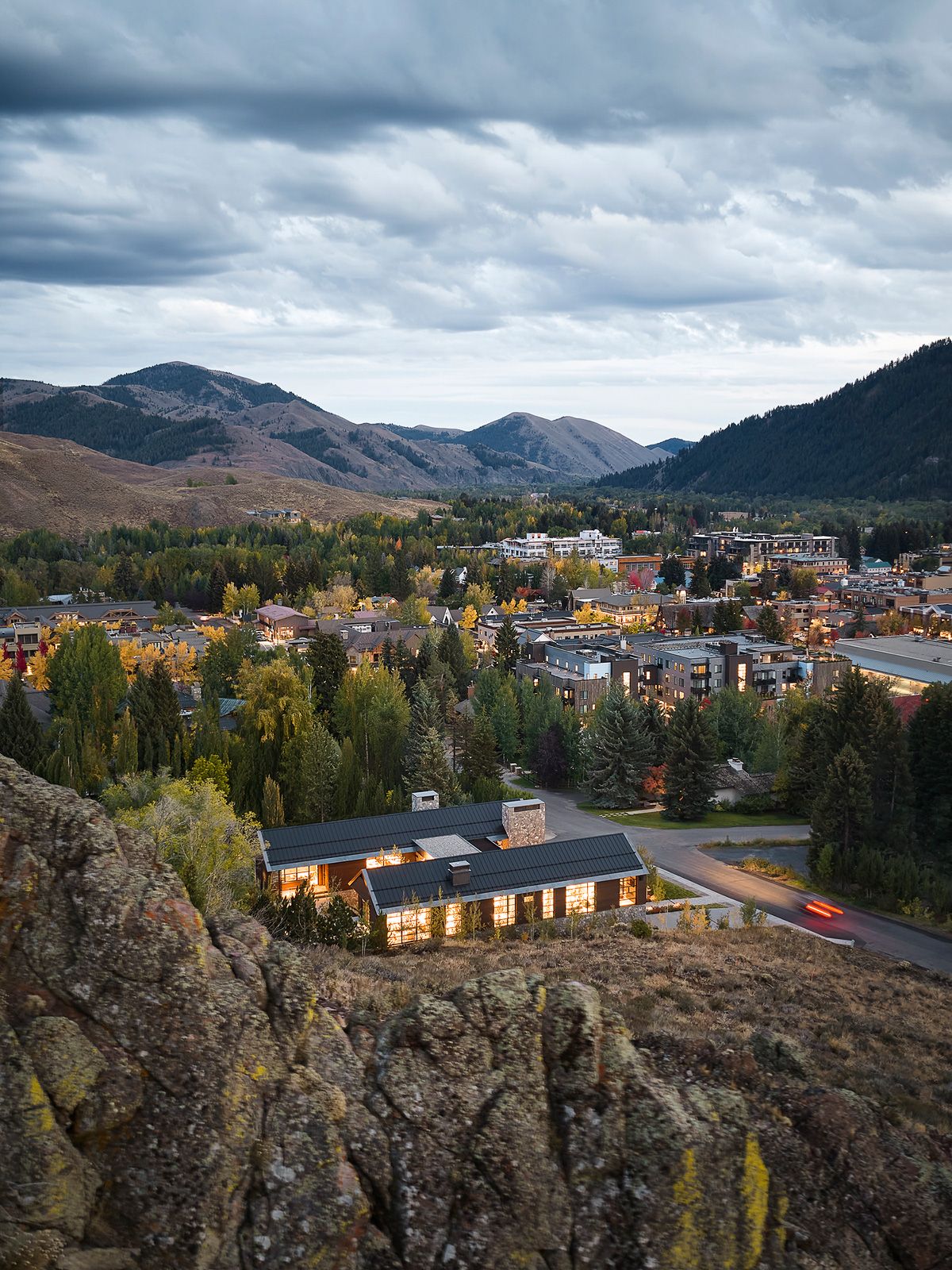 Modern mountain home exterior featuring a stone garage, large glass windows, and illuminated landscaping at twilight.
