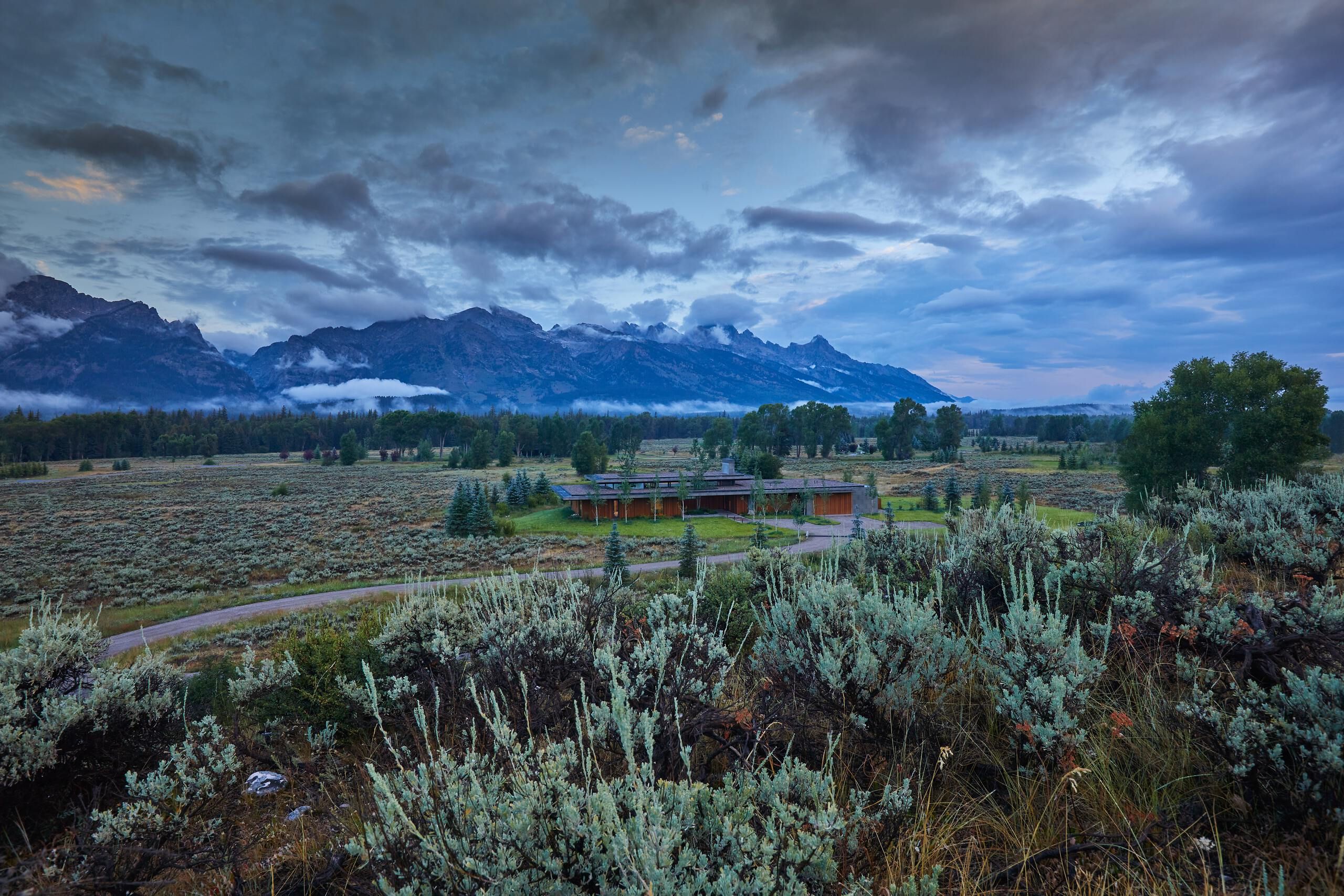 Dramatic view of the Grand Teton mountain range with cloud-covered peaks under a moody blue sky. A modern lodge is nestled in a vast valley of sagebrush with a winding dirt road leading to it, surrounded by distant evergreen forests.
