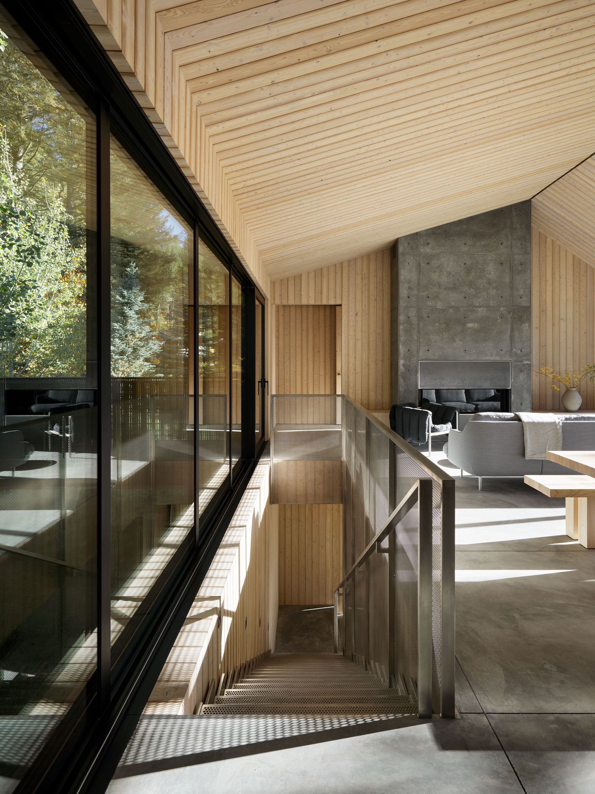 Modern, light-filled interior showcasing a living room with a concrete fireplace and a high vaulted wood ceiling. A perforated metal staircase descends alongside large floor-to-ceiling windows revealing outdoor trees.