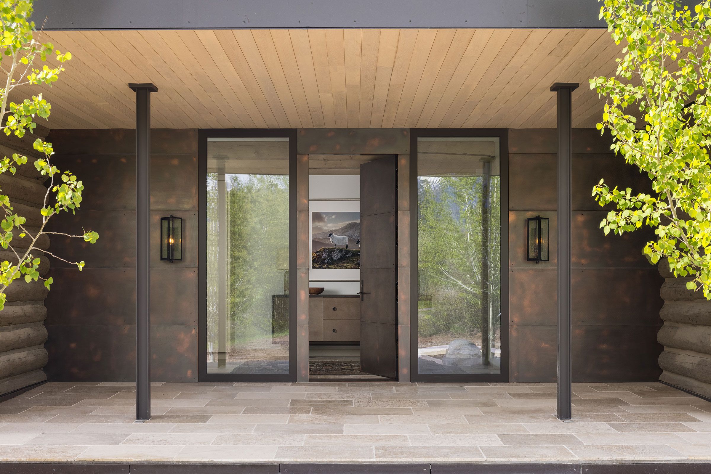 Exterior entryway of a modern rustic home featuring dark metal paneling, large glass windows, an open front door, and a stone tiled porch flanked by green trees.