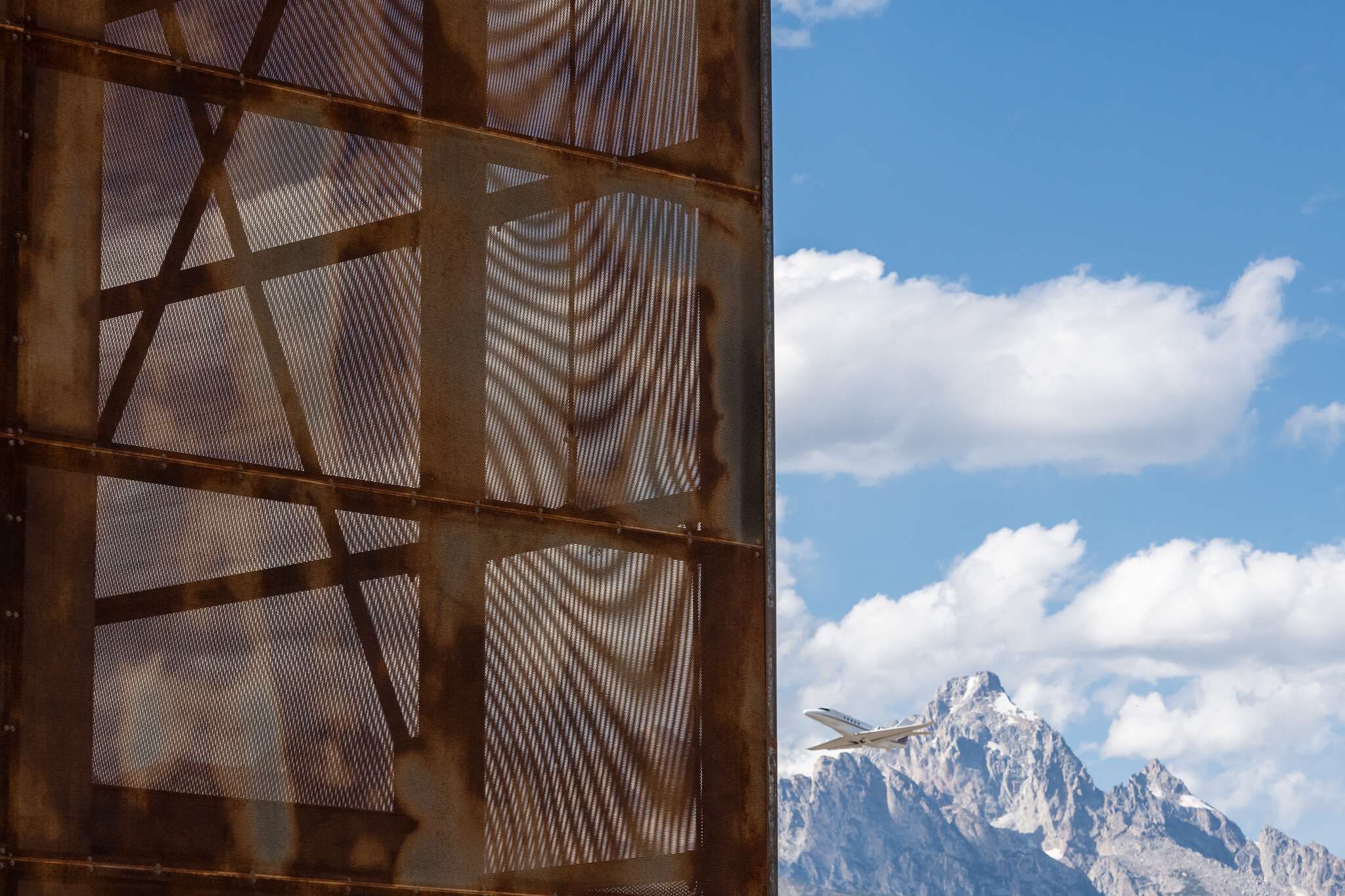 A rusted, perforated metal facade on the left, set against a bright blue sky with white clouds, snow-capped mountains, and a white private jet flying right.