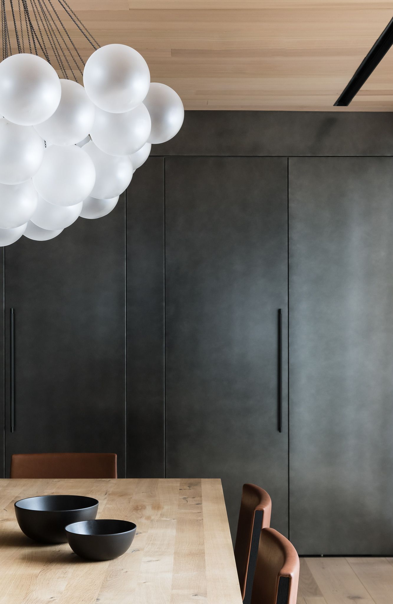 Modern dining room with a large spherical white chandelier, a light wood dining table with two matte black bowls, brown leather chairs, and a dark gray integrated cabinetry wall, complemented by wood plank ceiling and floor.