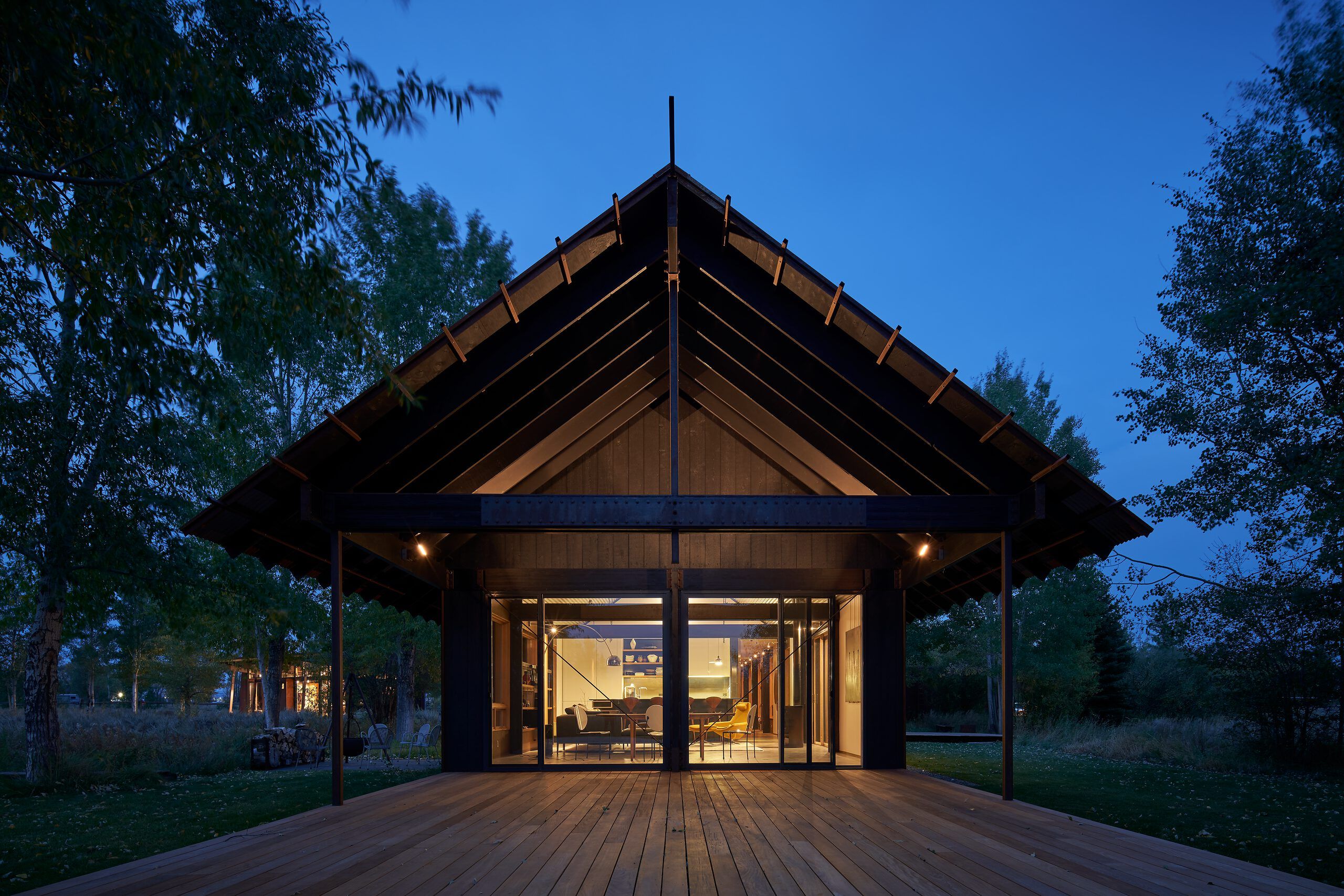 Modern gabled-roof home with a dark facade and exposed timber beams, brightly lit from within at twilight. A large wooden deck extends from its floor-to-ceiling glass windows, surrounded by a serene, tree-filled landscape under a deep blue sky.
