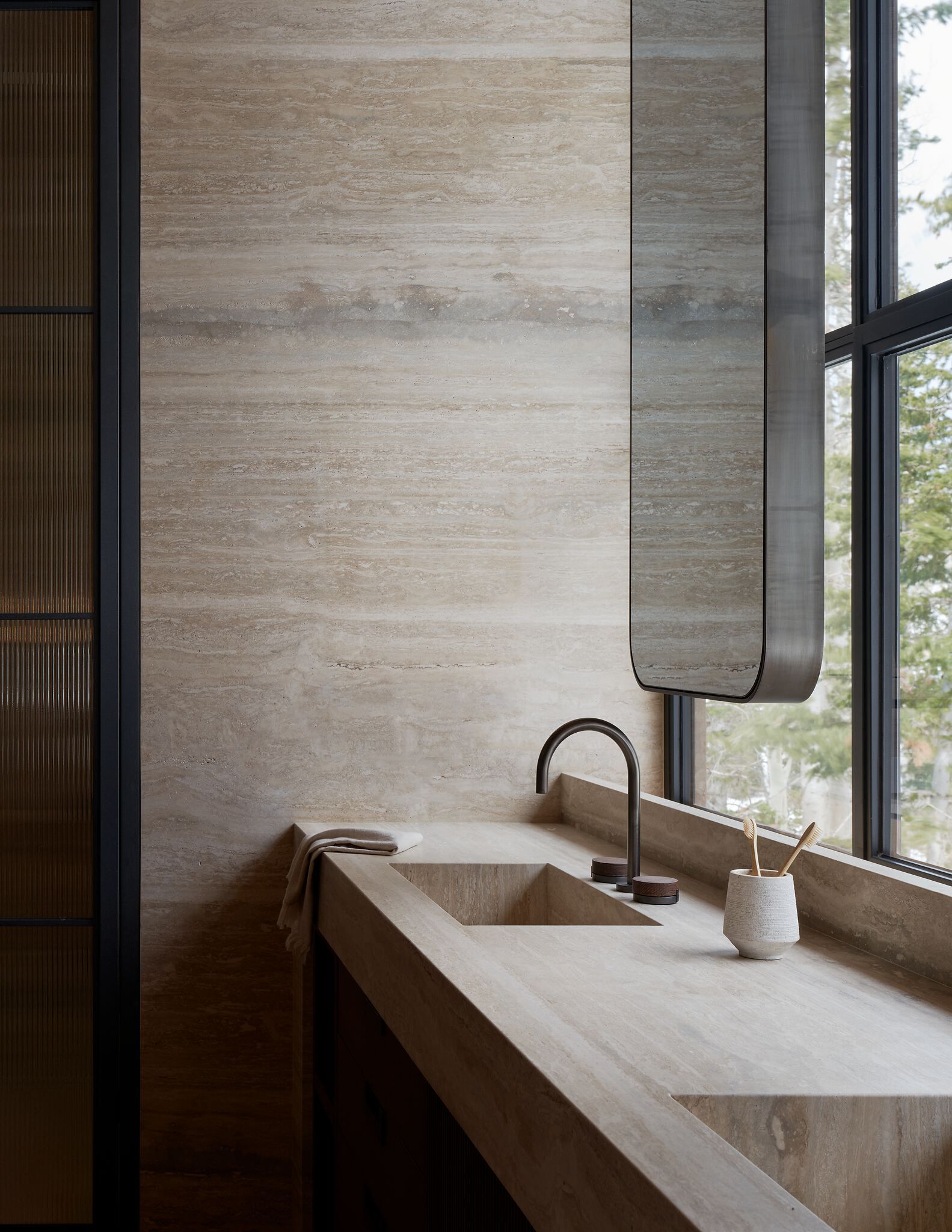 Modern minimalist bathroom with a beige travertine wall and integrated countertop sink. Features a dark curved faucet, a unique rounded mirror, and a large window offering a view of a winter forest.