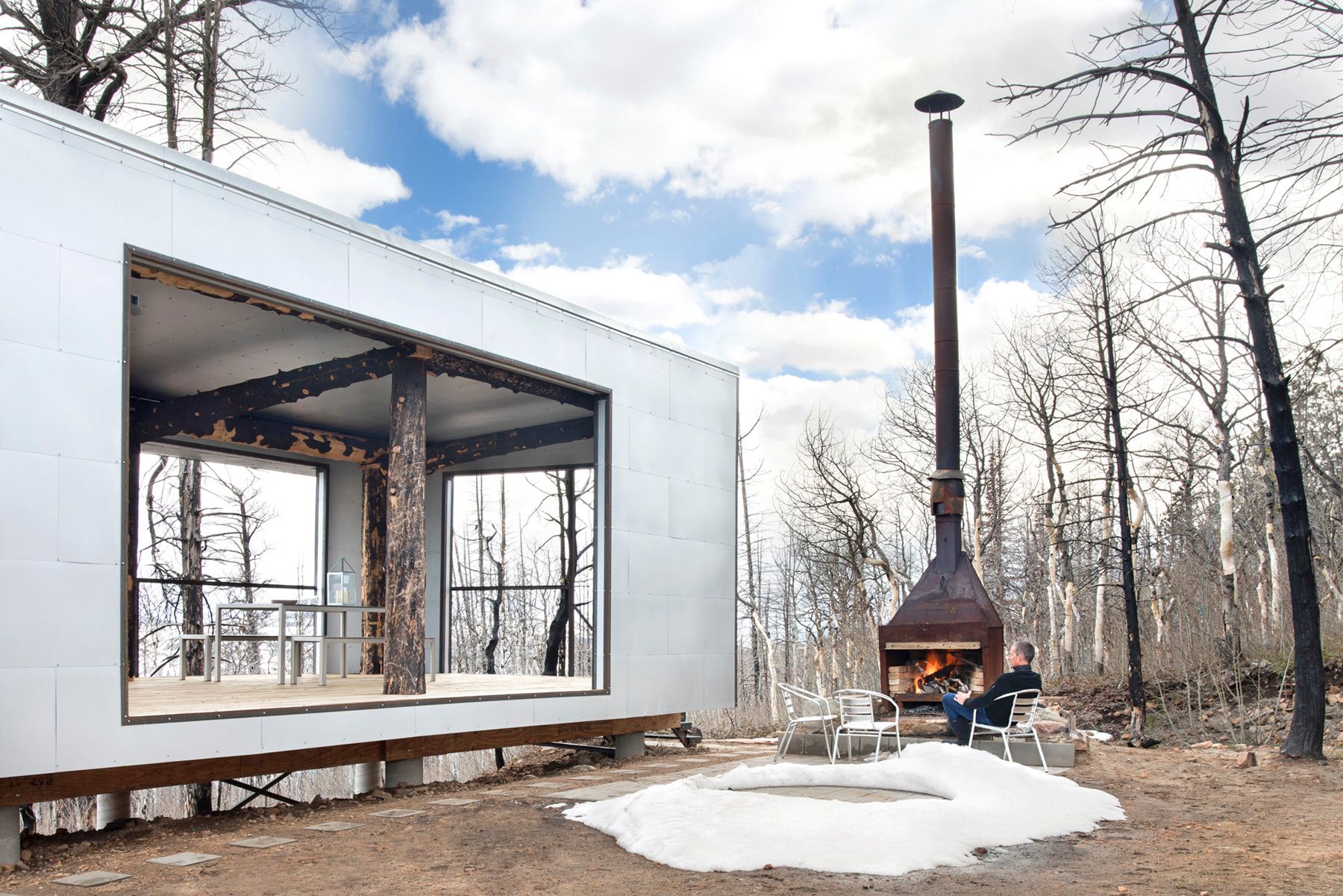 An exterior perspective of a modern, white modular cabin and its large, open patio. A man in black sits in a metal chair by a lit, tall metal outdoor fireplace, with a patch of snow on the ground and fire-damaged trees in the background.