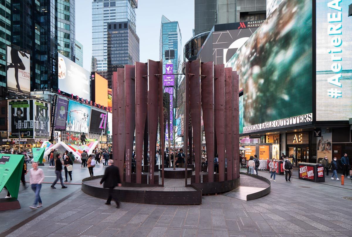 A bustling view of Times Square, New York City, featuring a reddish-brown cylindrical public art installation with a tree inside. Pedestrians move past giant illuminated billboards for stores like American Eagle Outfitters and Broadway shows, surrounded by tall skyscrapers.