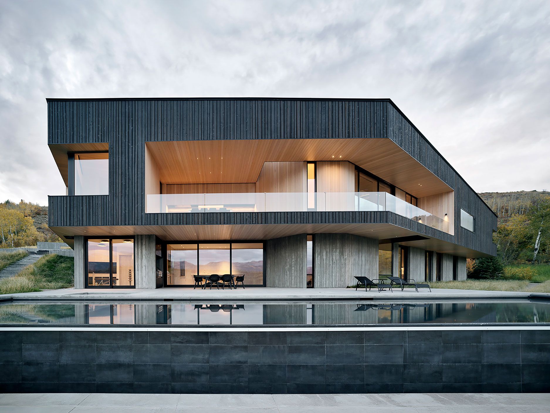 Modern two-story home with dark wood siding, concrete accents, upper-deck glass railing, and reflection pool.