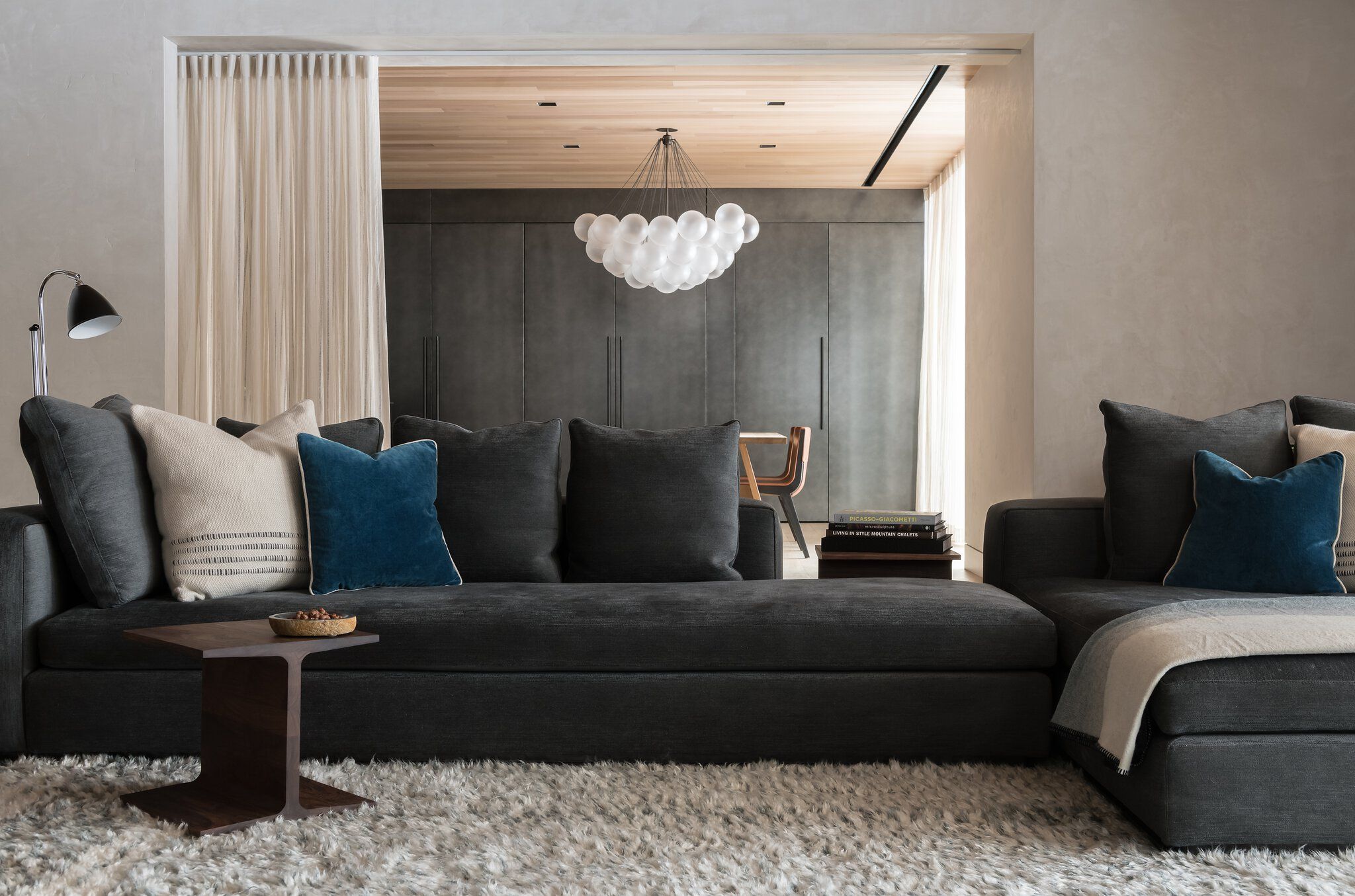 Modern open concept living room featuring a dark gray sectional sofa with cream and teal pillows on a plush white shag rug. An adjoining dining space reveals a wood-paneled ceiling, a sculptural bubble chandelier, and dark built-in cabinets.