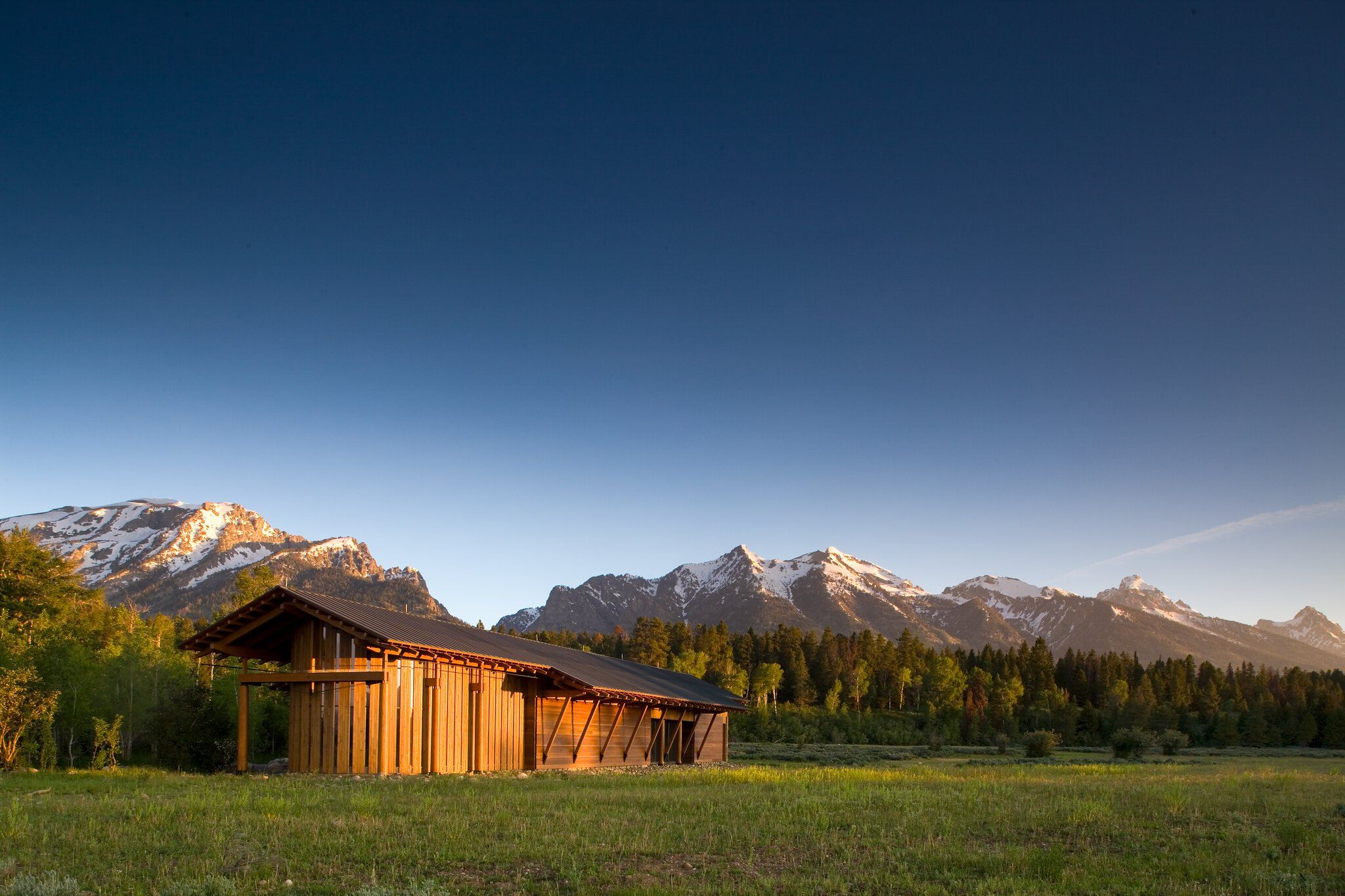 A rustic modern wooden building stands in a vibrant green meadow, with a dense forest and snow-capped mountain range in the background under a clear blue sky, illuminated by golden hour light.