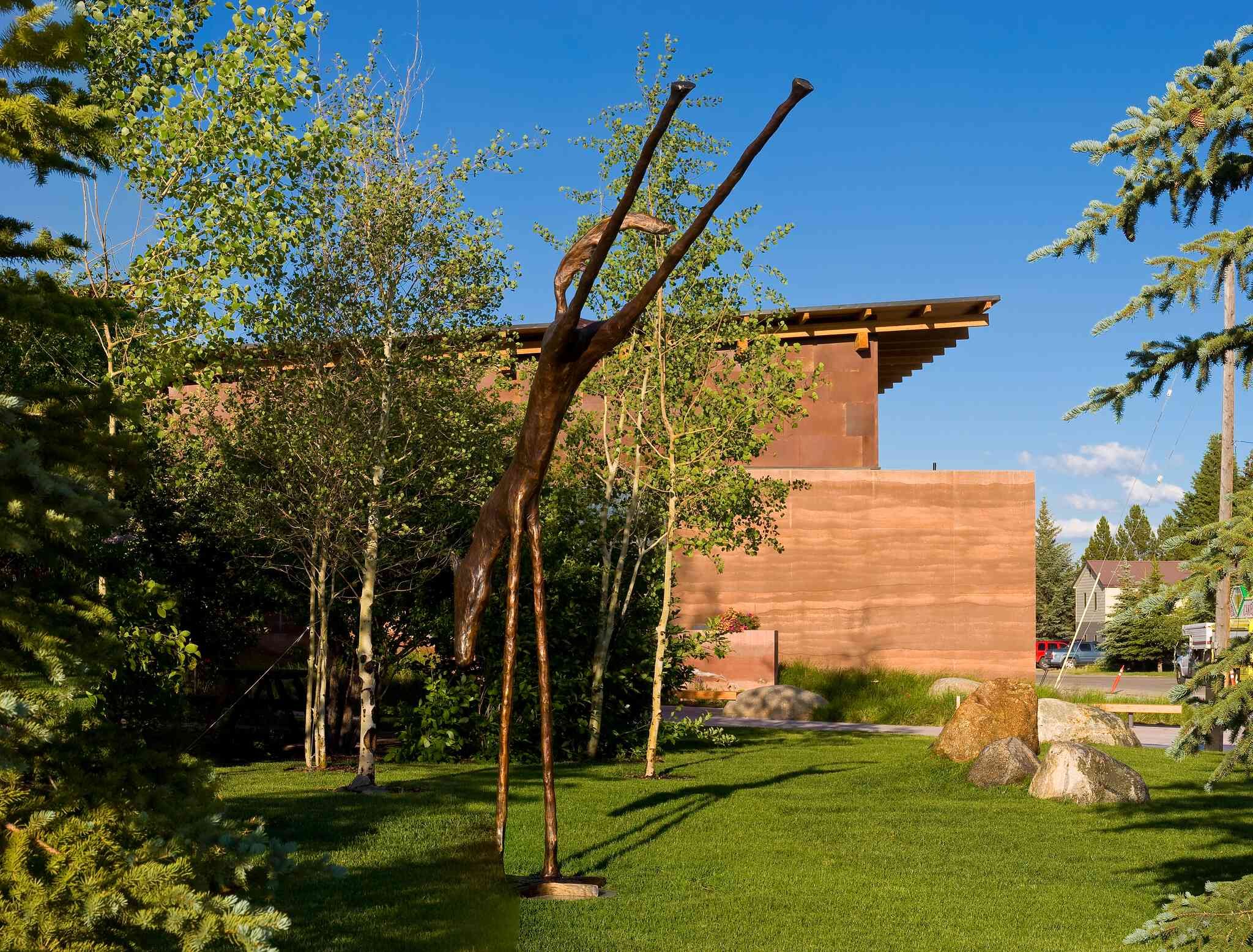 Tall, slender bronze sculpture of an upward-stretching figure on a lush green lawn, with a modern building featuring a layered, reddish-brown rammed earth wall and a sloped roof in the background, surrounded by trees and rocks under a blue sky.