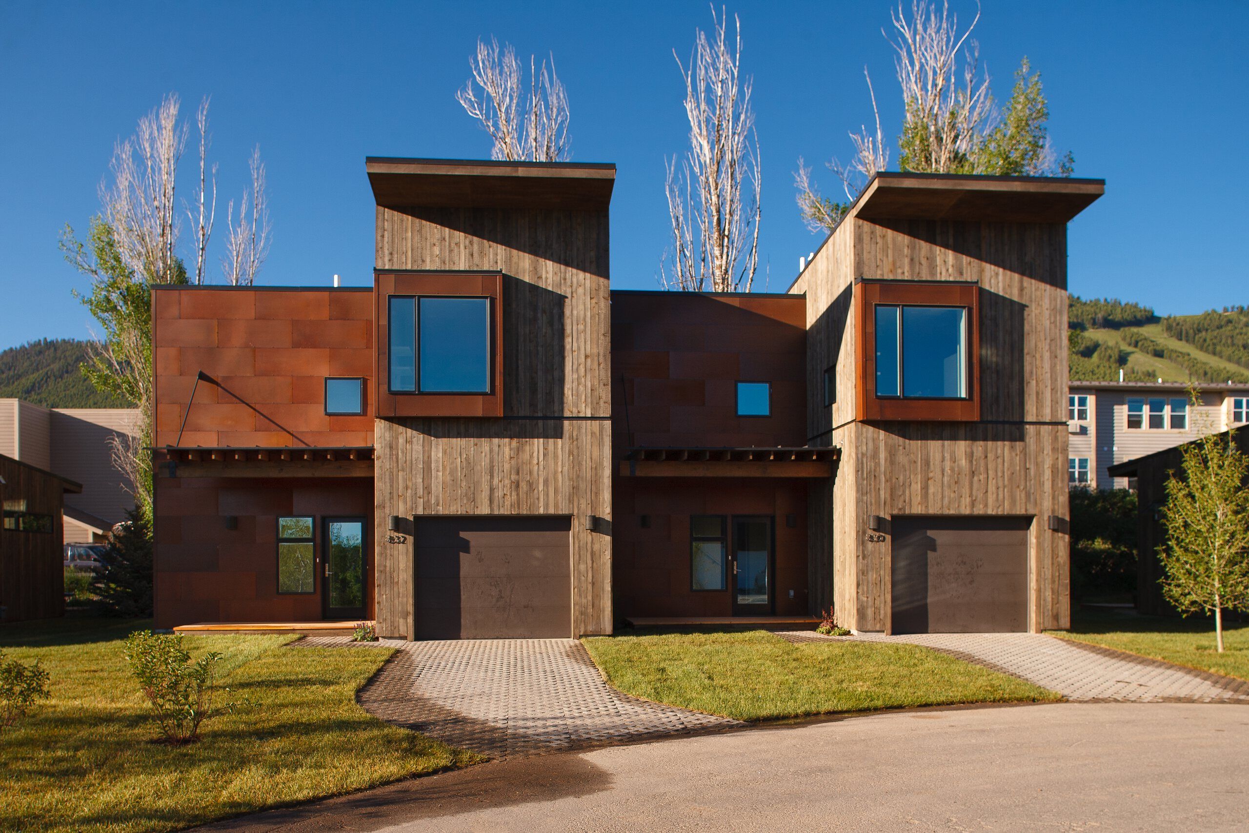 Two contemporary townhouses featuring a facade of rust-colored metal panels and vertical weathered wood siding under a bright blue sky. Green lawns and patterned paver driveways lead to dark garage doors. A tree-covered mountain with ski runs is visible in the background, suggesting a mountain resort setting.