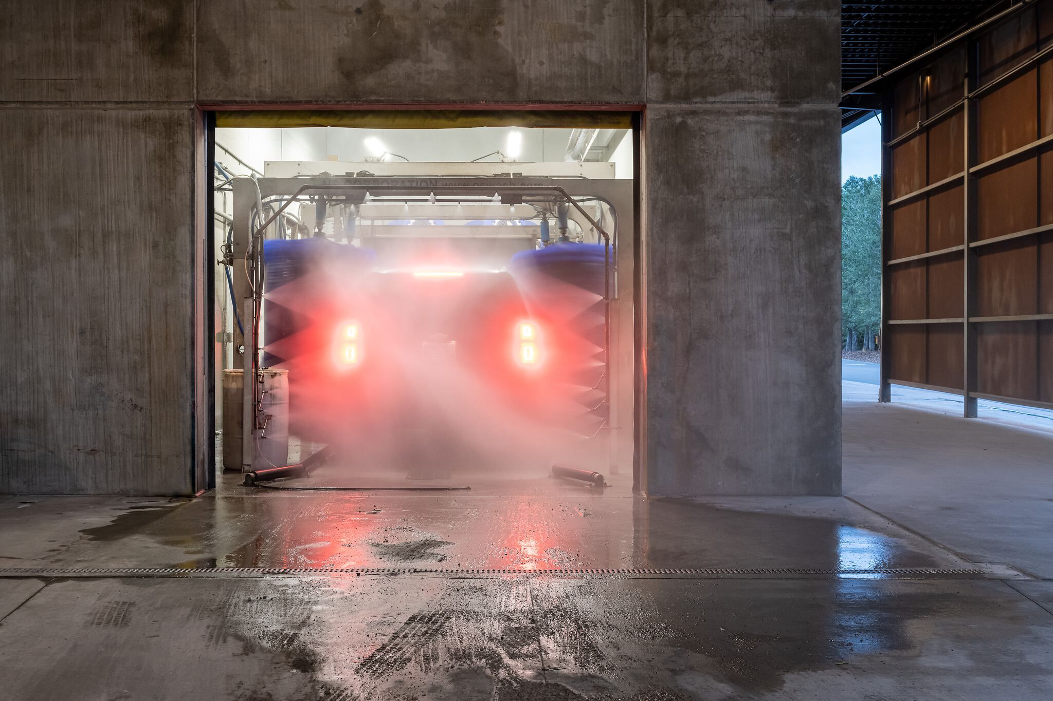 Automatic car wash tunnel interior, showing a car's rear with bright red taillights glowing through thick water spray and mist. Blue washing brushes are visible, framed by stark concrete walls and a wet concrete floor.