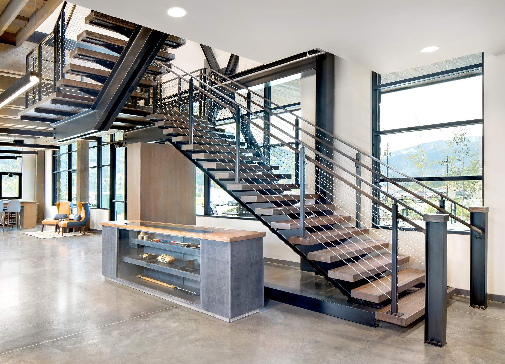 Modern industrial multi-level staircase featuring black steel stringers, floating wood treads, and stainless steel cable railings in a contemporary commercial lobby. The space has a polished concrete floor, large windows revealing a mountain and tree view, an exposed ceiling, and a sleek display counter with a wood top in the foreground.