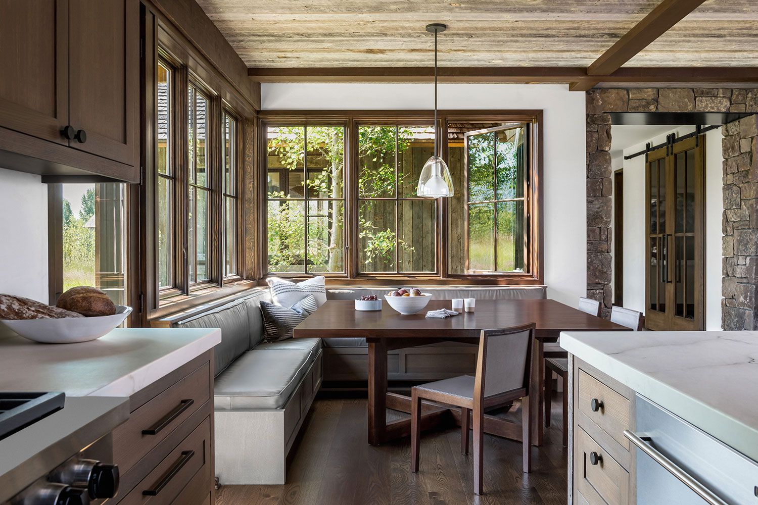 A cozy breakfast nook next to a kitchen island, featuring a built-in grey banquette, a dark wood dining table, and large windows overlooking a lush green yard.