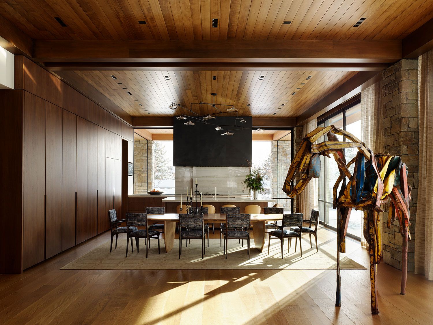 A spacious modern dining room with a long wood table, geometric chairs, wood-planked ceiling, and a large abstract horse sculpture made of colorful scrap metal.