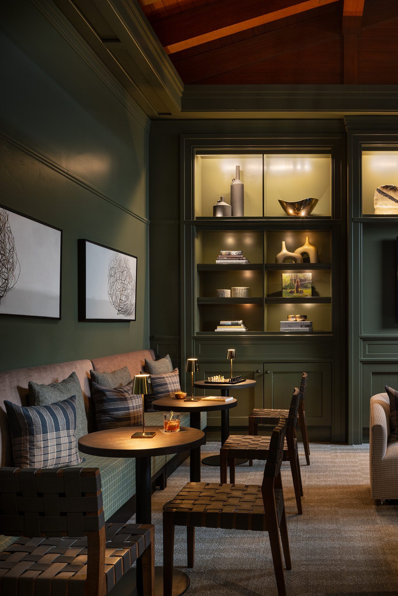 A cozy, sophisticated lounge featuring dark green paneled walls, built-in illuminated bookshelves, plush banquette seating with plaid pillows, and woven leather chairs around small tables, one holding a chessboard.
