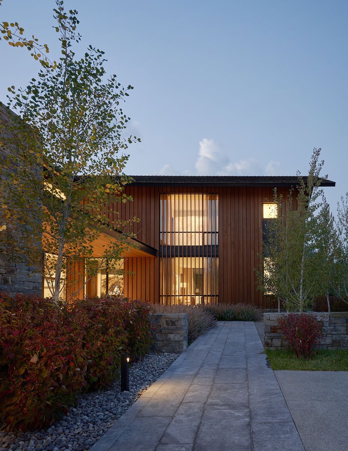 A stone walkway leading toward a modern house with vertical wood siding and glowing floor-to-ceiling windows at evening, framed by autumn landscaping.