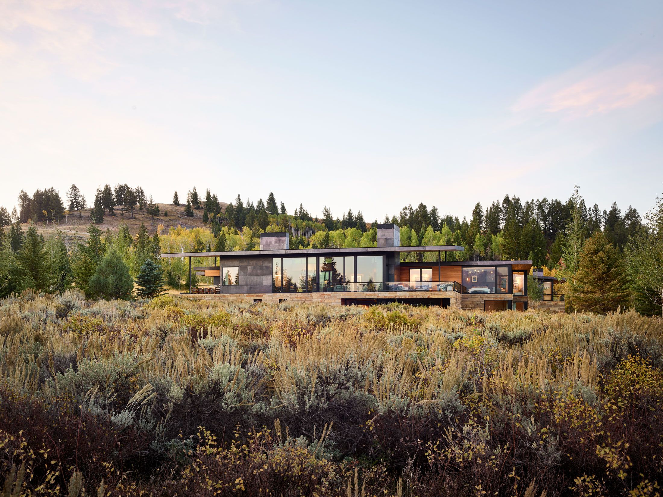 Modern mountain home featuring expansive glass windows and a stone and wood exterior, nestled in a golden autumn meadow. Evergreen and yellow-leaved trees line the distant hills under a soft evening sky.