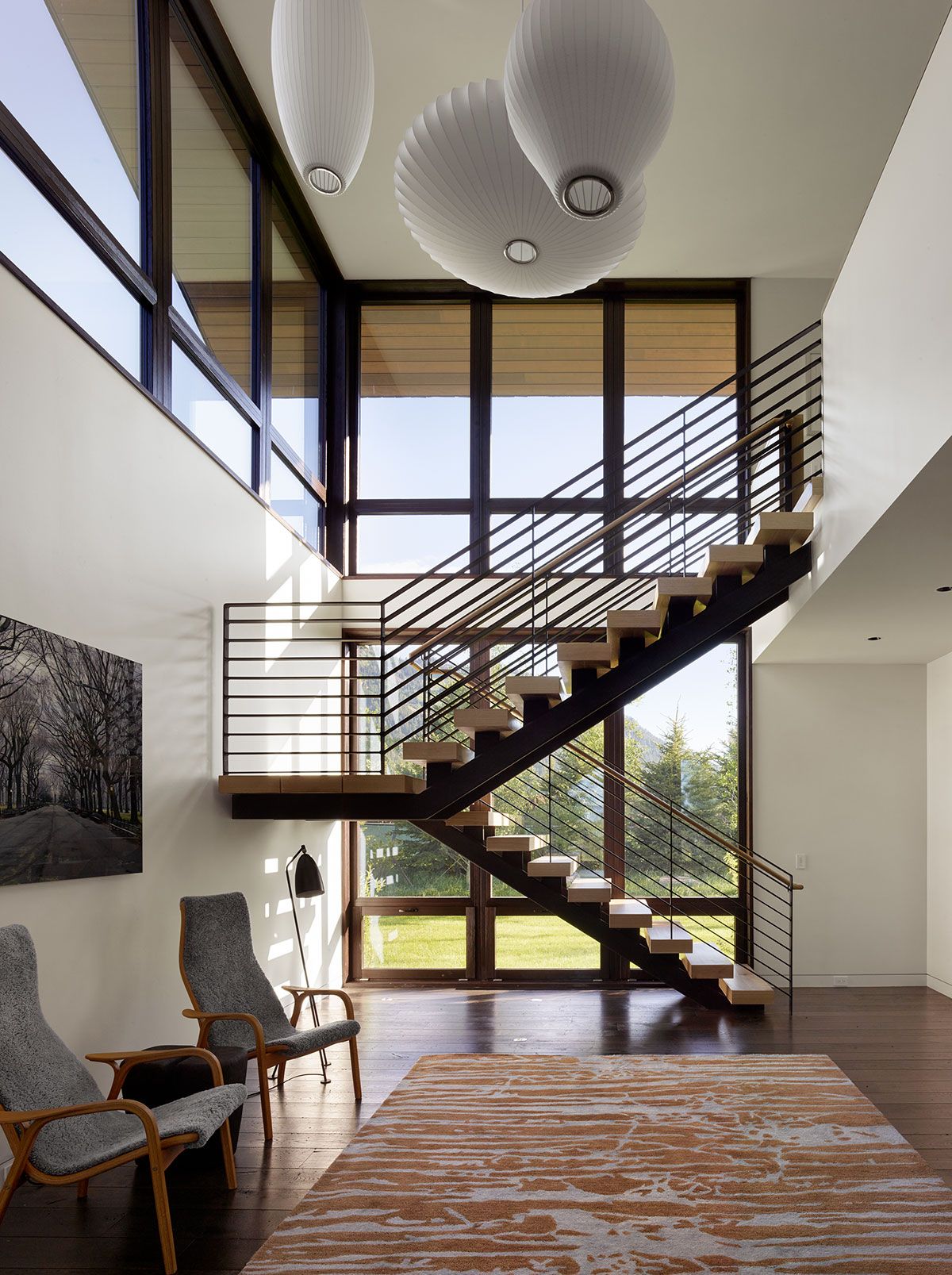 A bright, two-story entryway featuring a black steel floating staircase with light wood treads, large grid windows, mid-century modern armchairs, and hanging cocoon pendant lights.