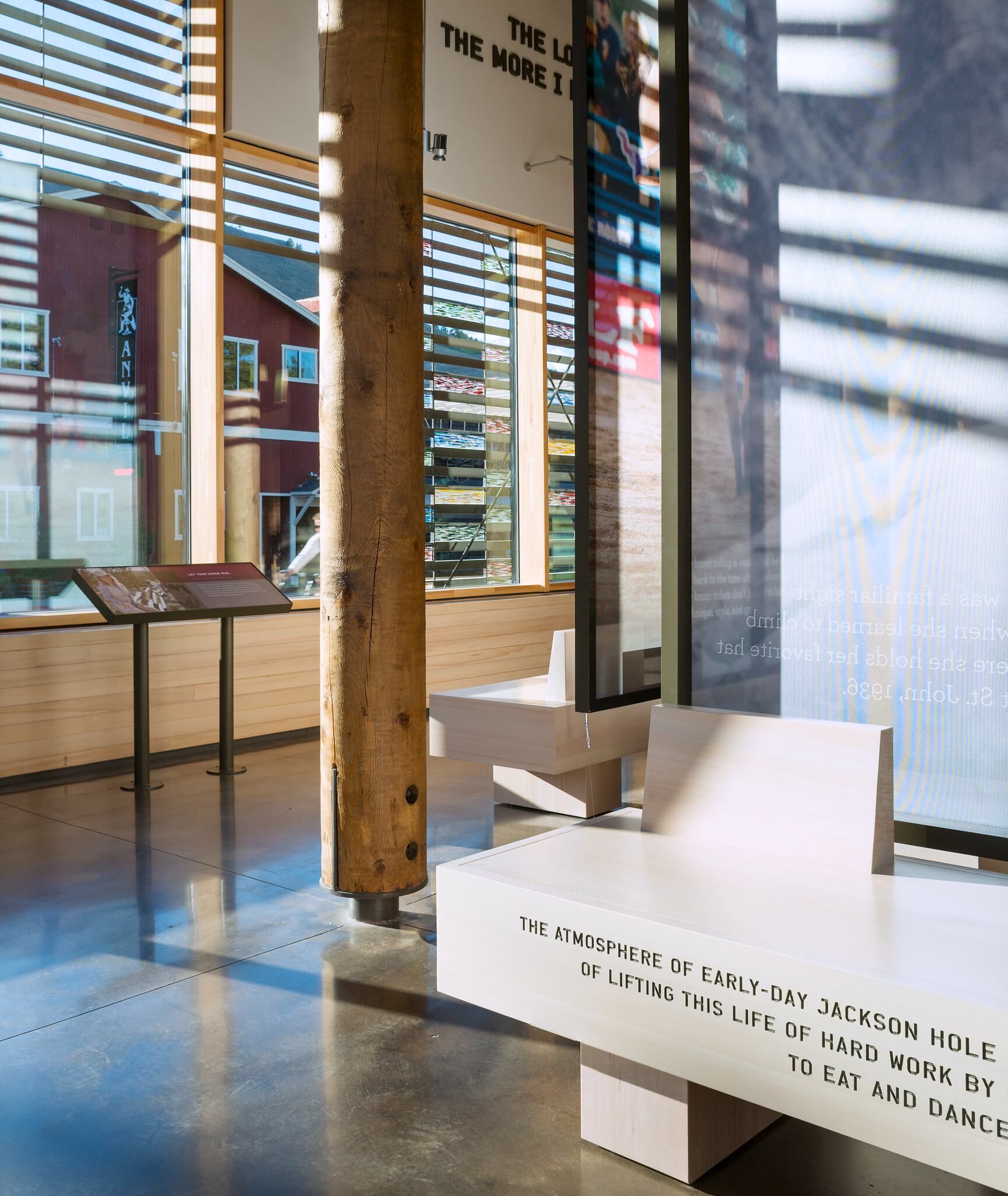 Modern museum or visitor center interior with large windows and horizontal blinds, showcasing a rustic wooden pillar and polished concrete floor. Light wooden benches are inscribed with text about early-day Jackson Hole, alongside transparent digital display panels and an information stand with historical photos. Sunlight streams in, casting shadows, and reflecting a red building with an Anvil Hotel sign in the windows.