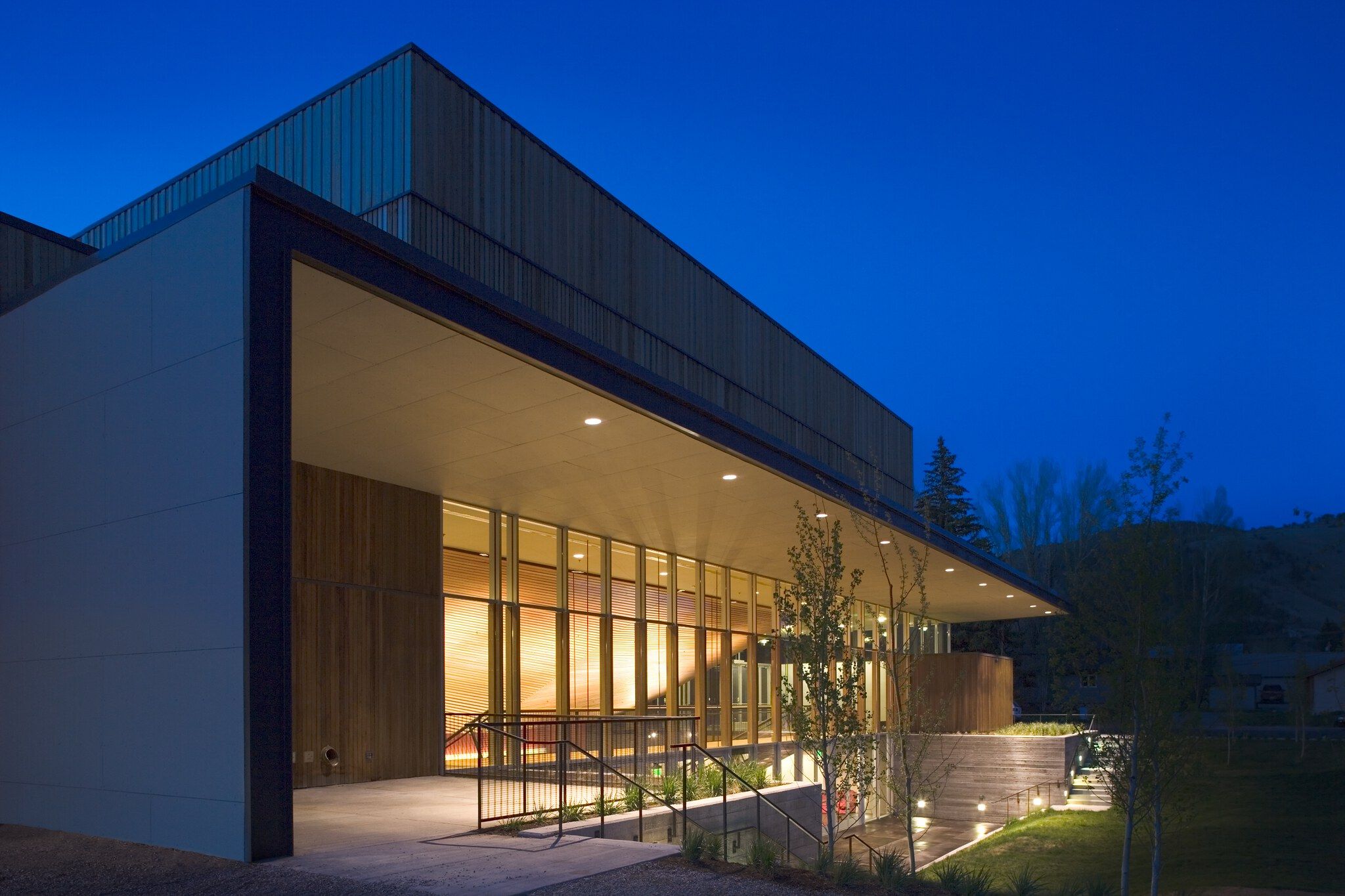 Contemporary building illuminated at twilight, featuring a wood and glass facade under an overhanging canopy with recessed lights. An illuminated pathway with steps and railings leads to the entrance, set against a deep blue evening sky and natural landscape.