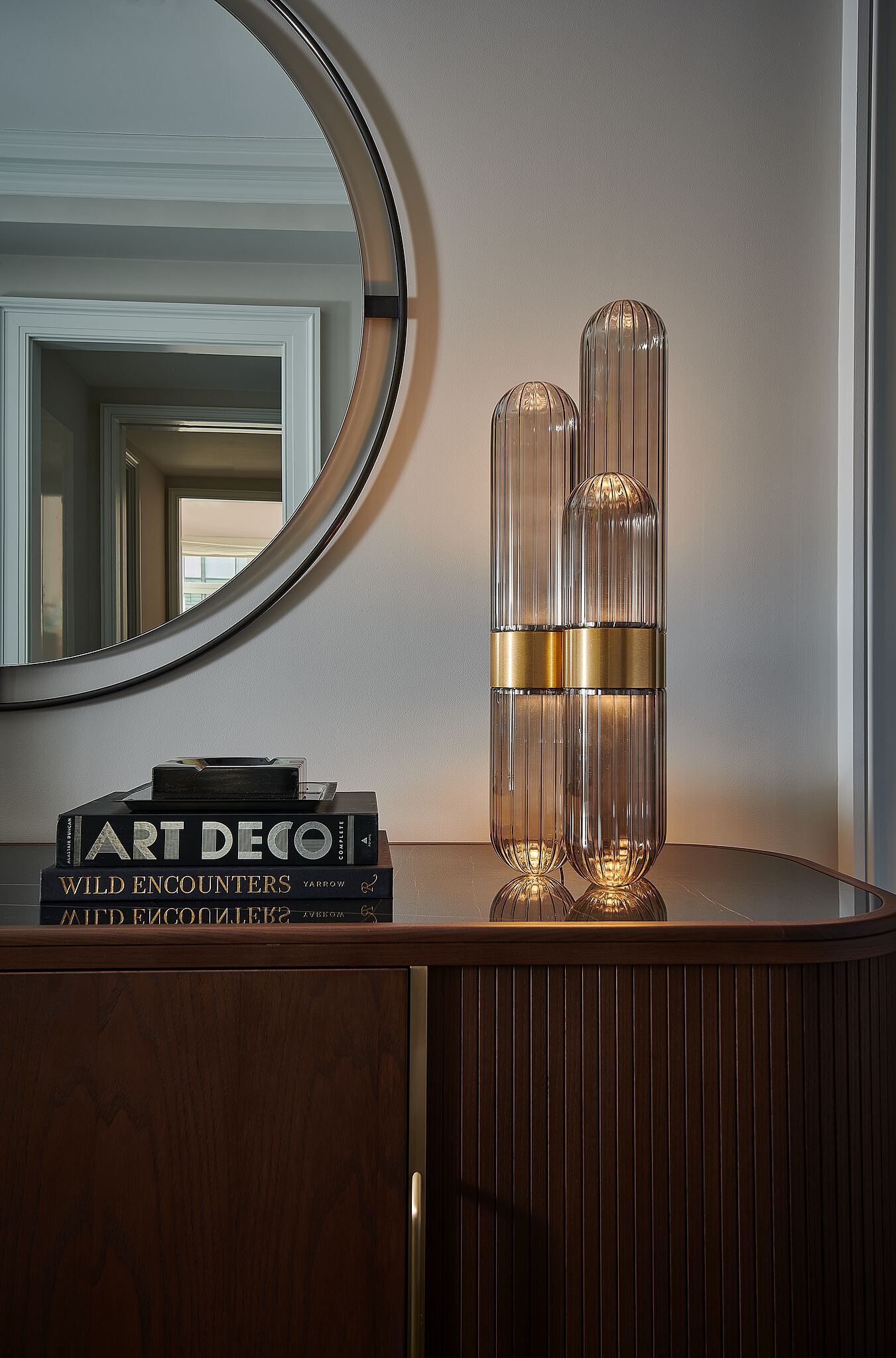 Elegant entryway featuring a large round mirror above a dark wood console cabinet with fluted details and brass hardware. On its polished surface sit three glowing ribbed glass lamps with a brass accent band, alongside a stack of decor books including 'ART DECO'.