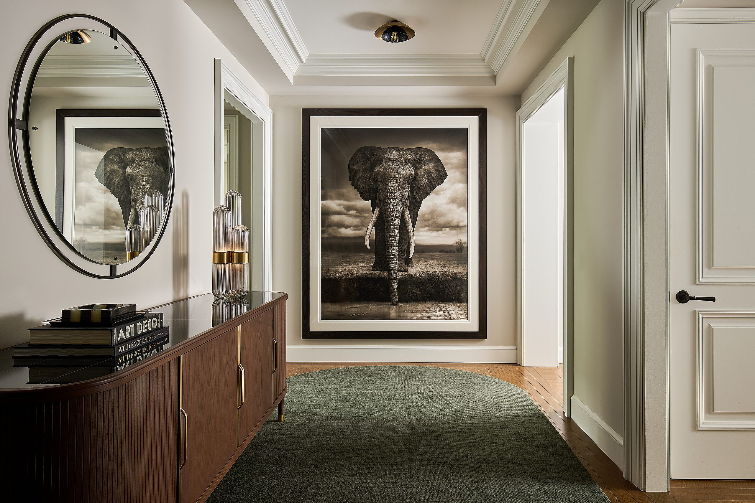 Elegant entryway featuring a large framed black and white elephant artwork, a dark wood console table with fluted glass sculptures and design books, and an oval mirror reflecting the art. A dark green rug covers part of the light hardwood floor, leading towards a paneled white door.