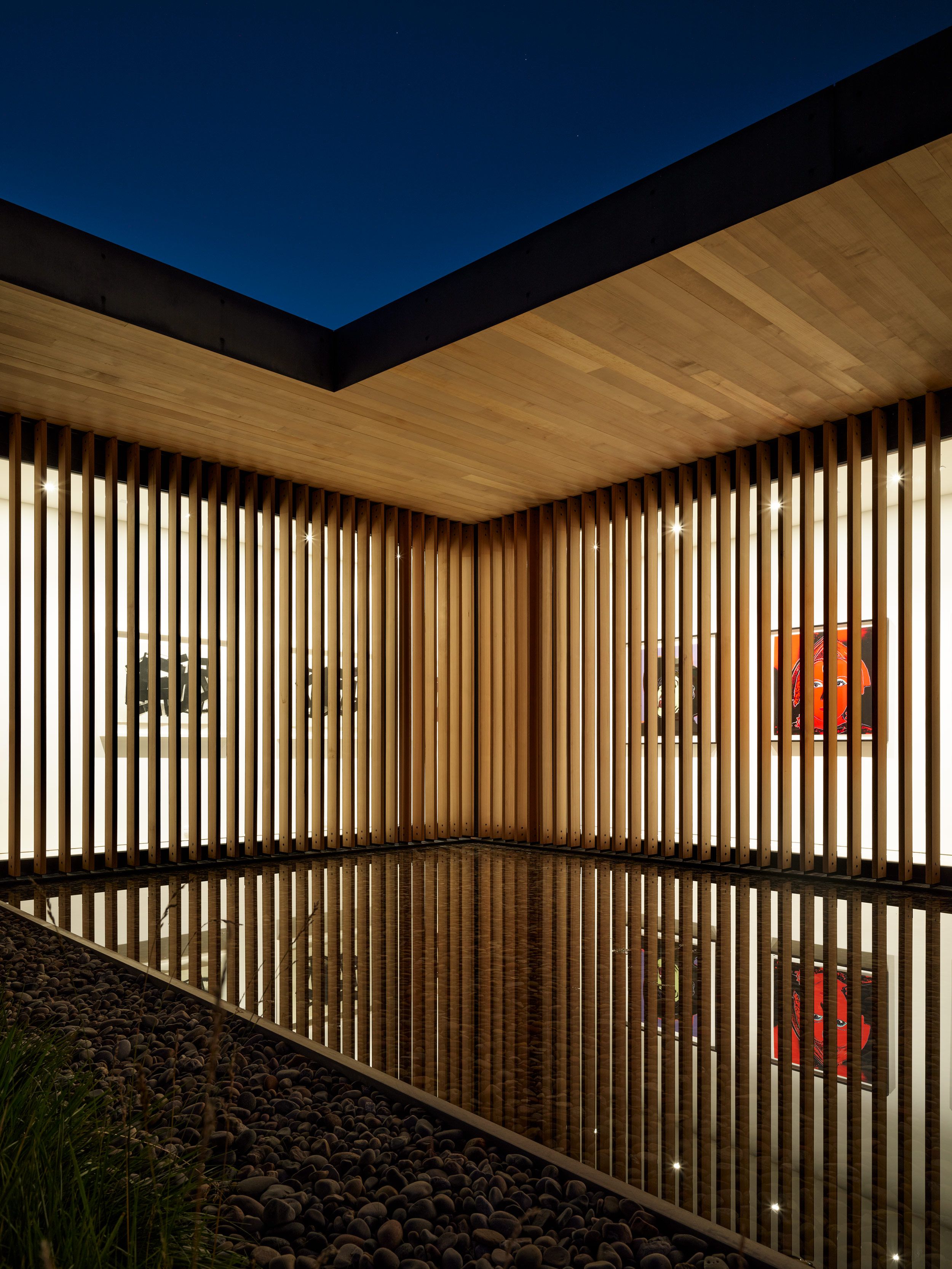 Modern architectural pavilion at night, featuring vertical wooden slatted walls behind which an illuminated art gallery displays colorful pop art. A serene reflecting pool mirrors the structure below a dark blue sky, while dark river pebbles and sparse greenery frame the bottom left.