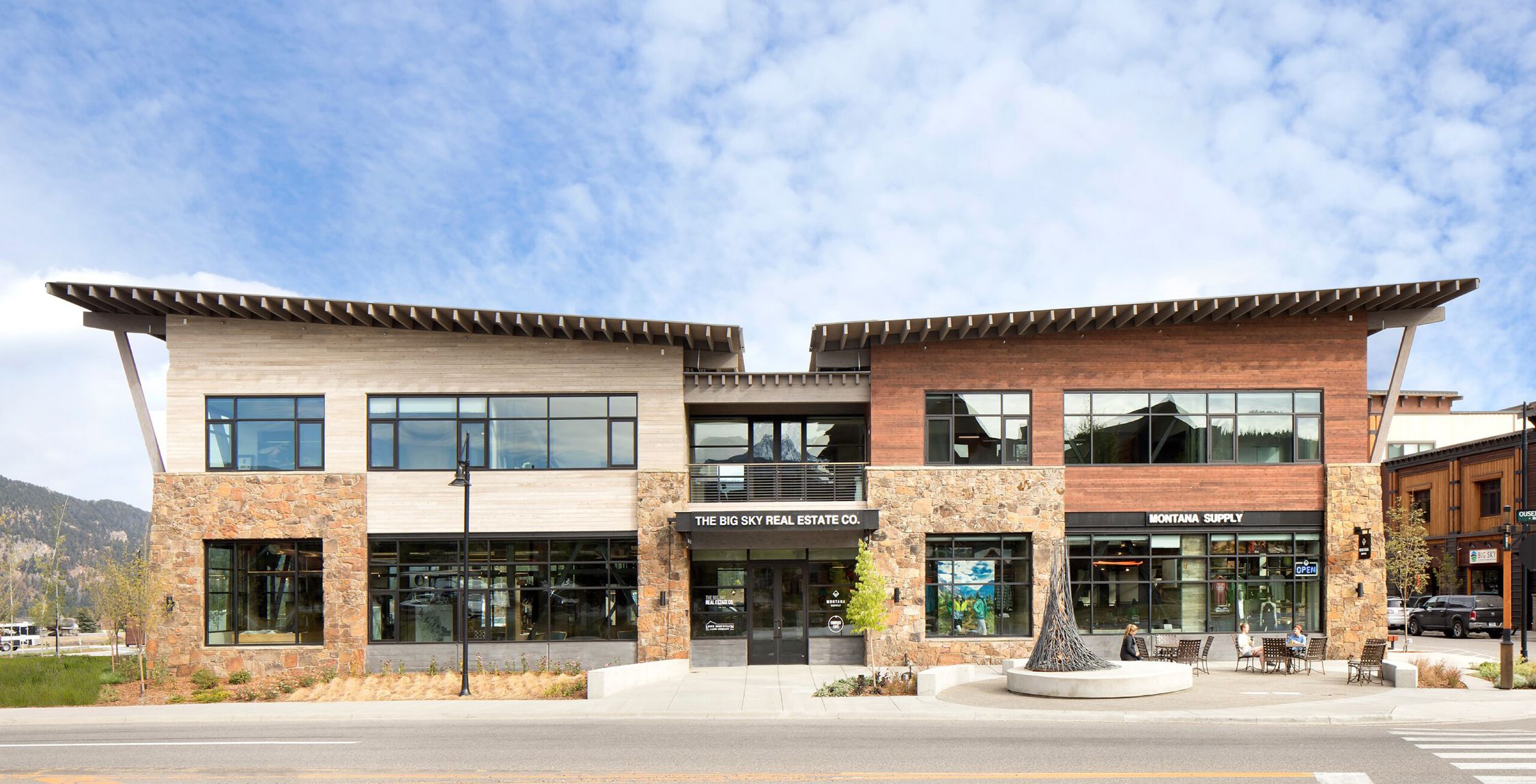Modern commercial building in Big Sky, Montana, featuring a stone and wood facade. It houses The Big Sky Real Estate Co. and Montana Supply, with people seated at outdoor tables on the sidewalk under a blue sky with scattered clouds and distant mountains.