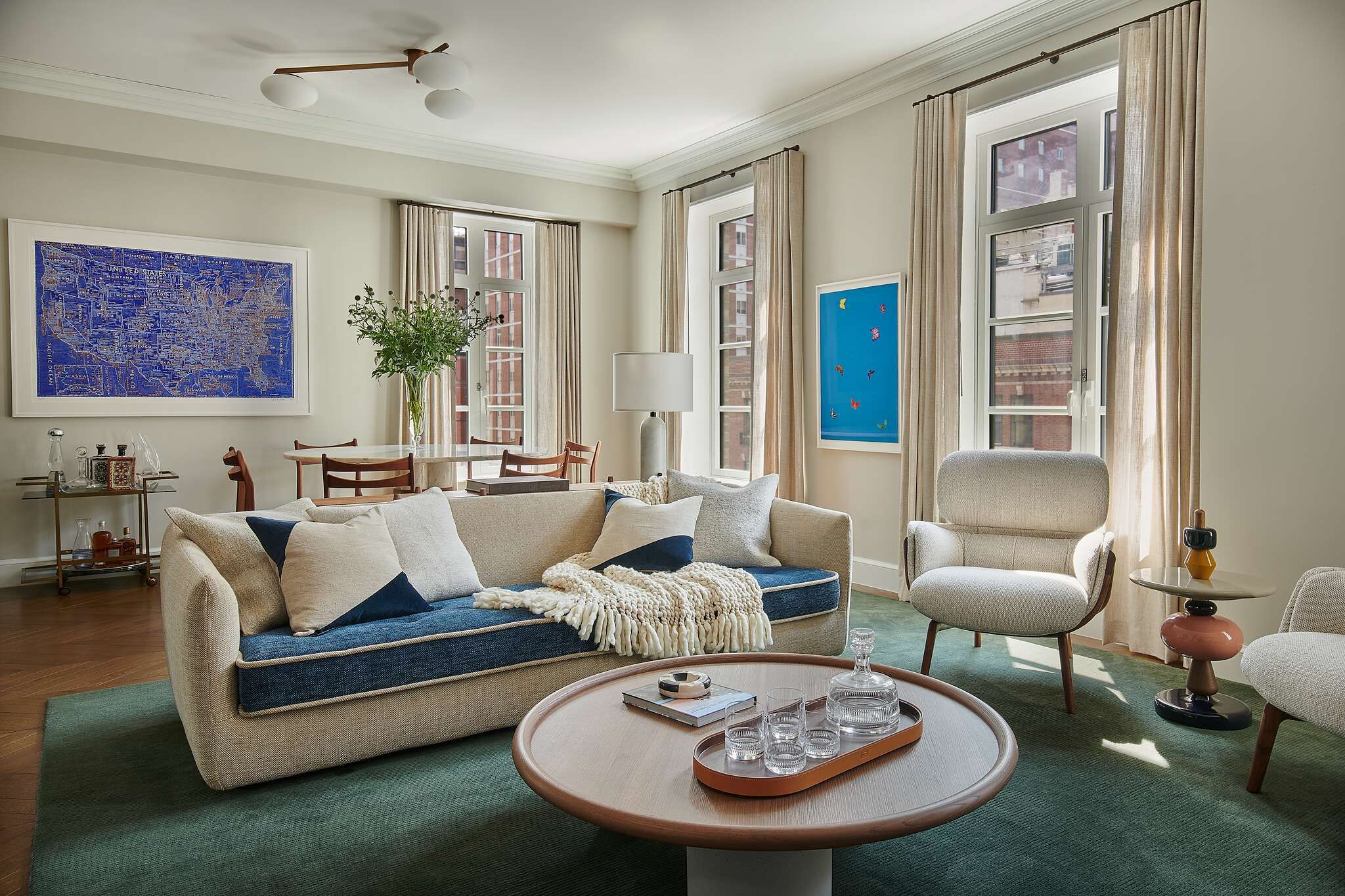 Elegant modern living room featuring a beige sofa with deep blue cushions and accent pillows, a round wood coffee table on a green ribbed area rug, and light-colored upholstered armchairs. Tall windows with neutral drapes bring in natural light, complemented by a large framed blue map artwork and a smaller blue piece with colorful butterflies on the walls.