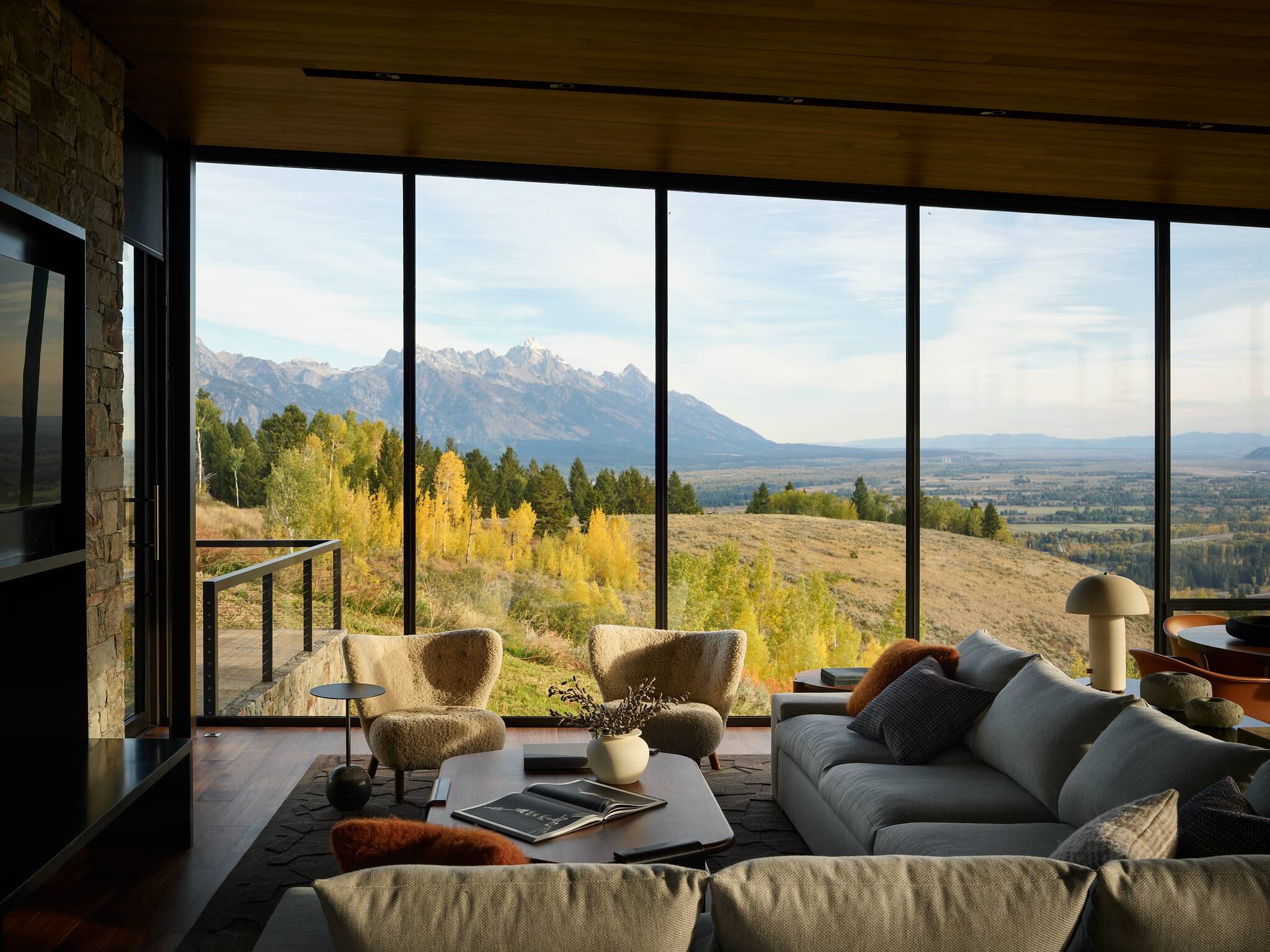 Luxurious modern living room with floor-to-ceiling windows overlooking the Grand Teton mountains and a vibrant autumn landscape. Features a plush grey sectional sofa, two textured armchairs, a wooden coffee table, and a warm wooden ceiling.