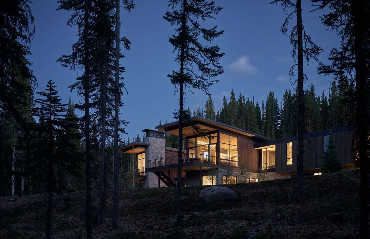 Luxury modern home featuring wood siding, stone accents, and expansive windows with warm interior lighting, nestled on a forested hillside at twilight.
