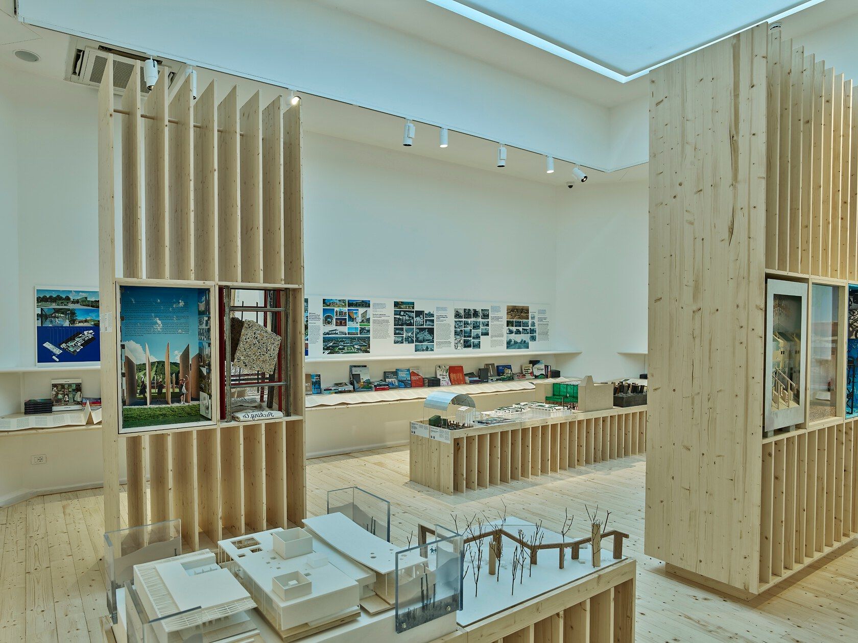 Modern architectural exhibition interior showcasing white building and landscape models on light wooden display tables. The space features tall, slatted wooden partitions with embedded displays, and project posters and diagrams line the white walls. Light wood flooring completes the bright, clean design.