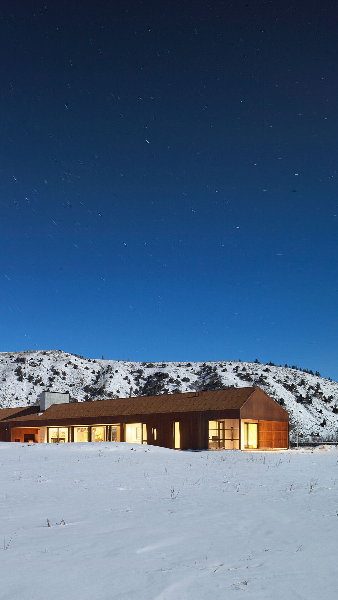 Modern corten steel house with warmly lit windows in a snowy winter landscape. Star trails streak across a deep blue night sky above a snow-dusted mountain.