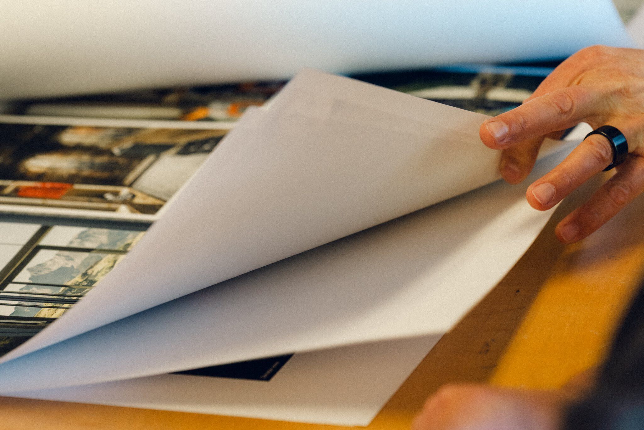 A hand with a black ring turns the thick white pages of a large photo portfolio, revealing blurred images of landscapes and architecture in the background.