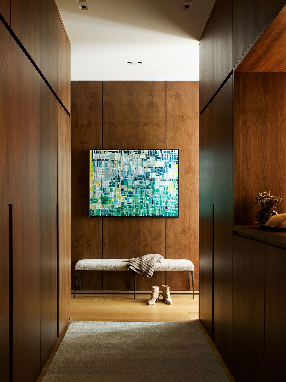 A modern wood-paneled hallway featuring a vibrant blue and green abstract painting hanging above a upholstered white bench with shoes underneath.